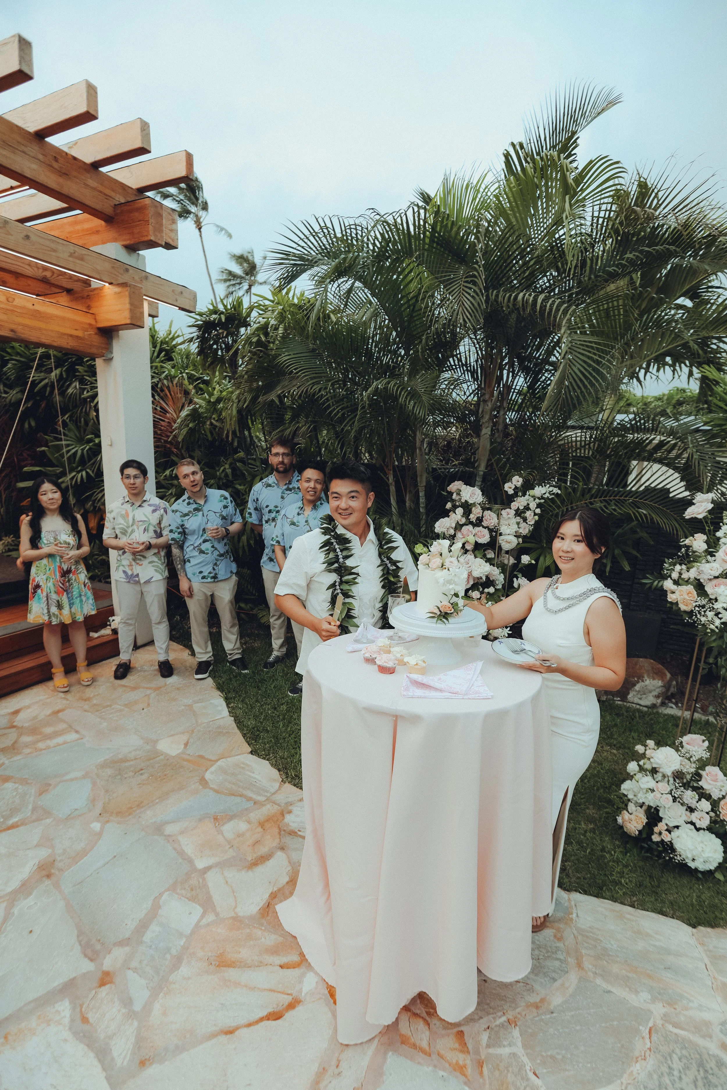 Couple standing beside a wedding cake with guests gathered in a tropical outdoor setting.
