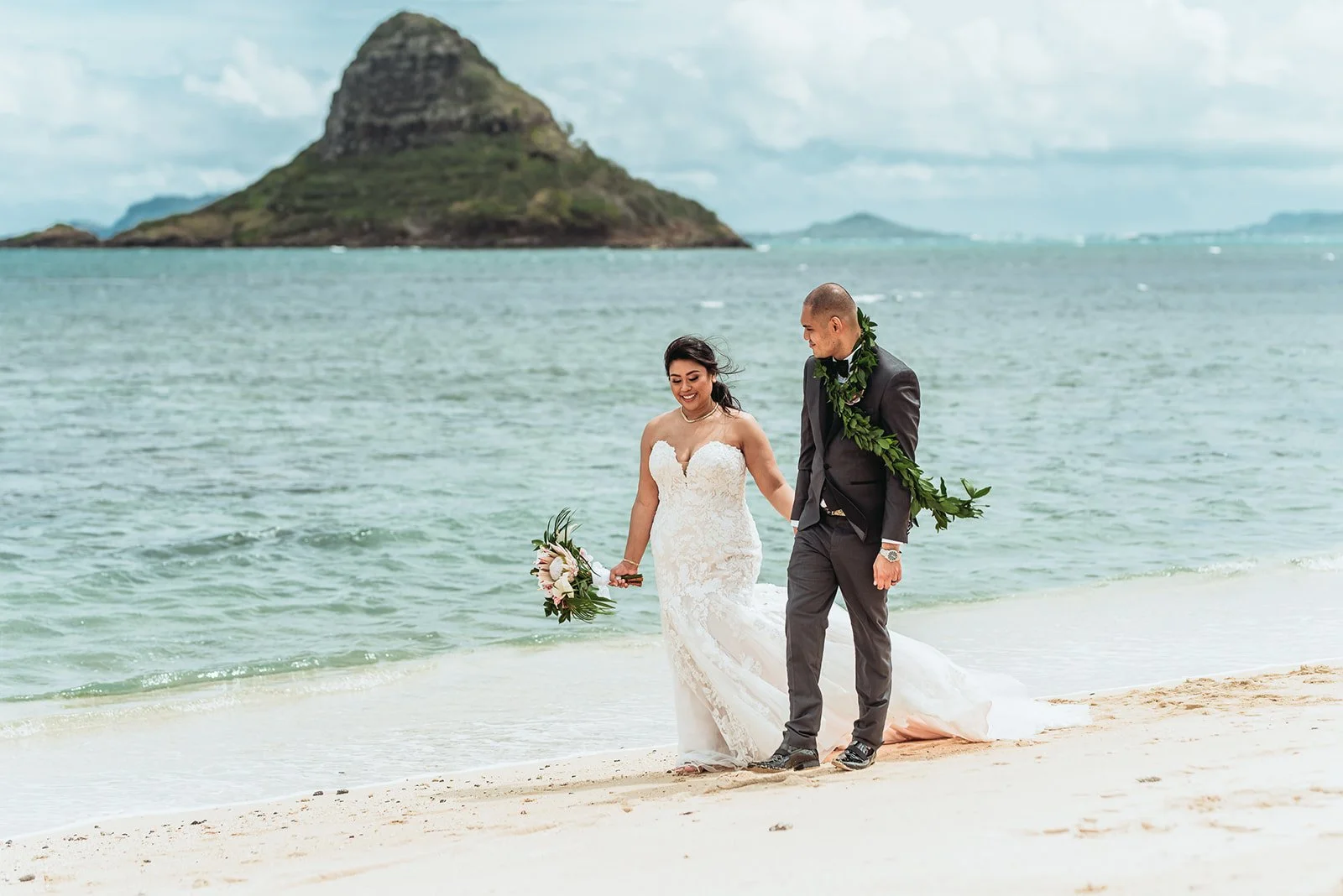 Bride and groom walking together along the shoreline with ocean and mountain views behind them.