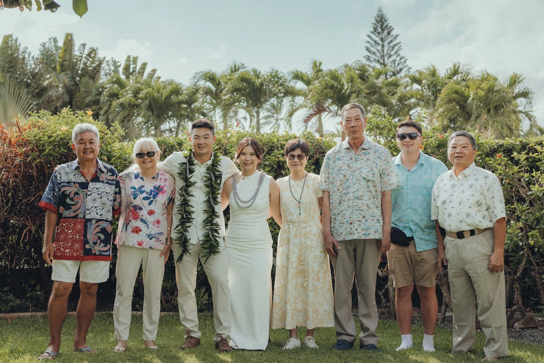 Emma and Evan in white, posing outdoors with loved ones in aloha attire.