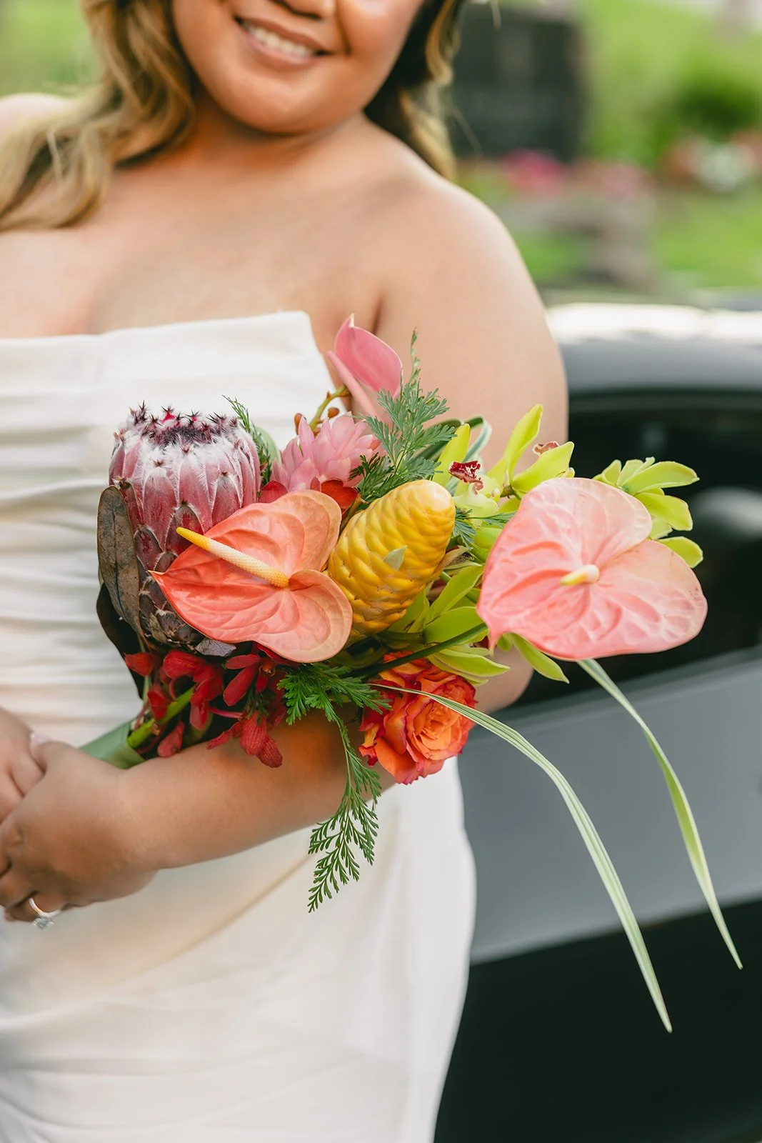Bride holding a colorful tropical bouquet with pink anthuriums, orchids, roses, and greenery.