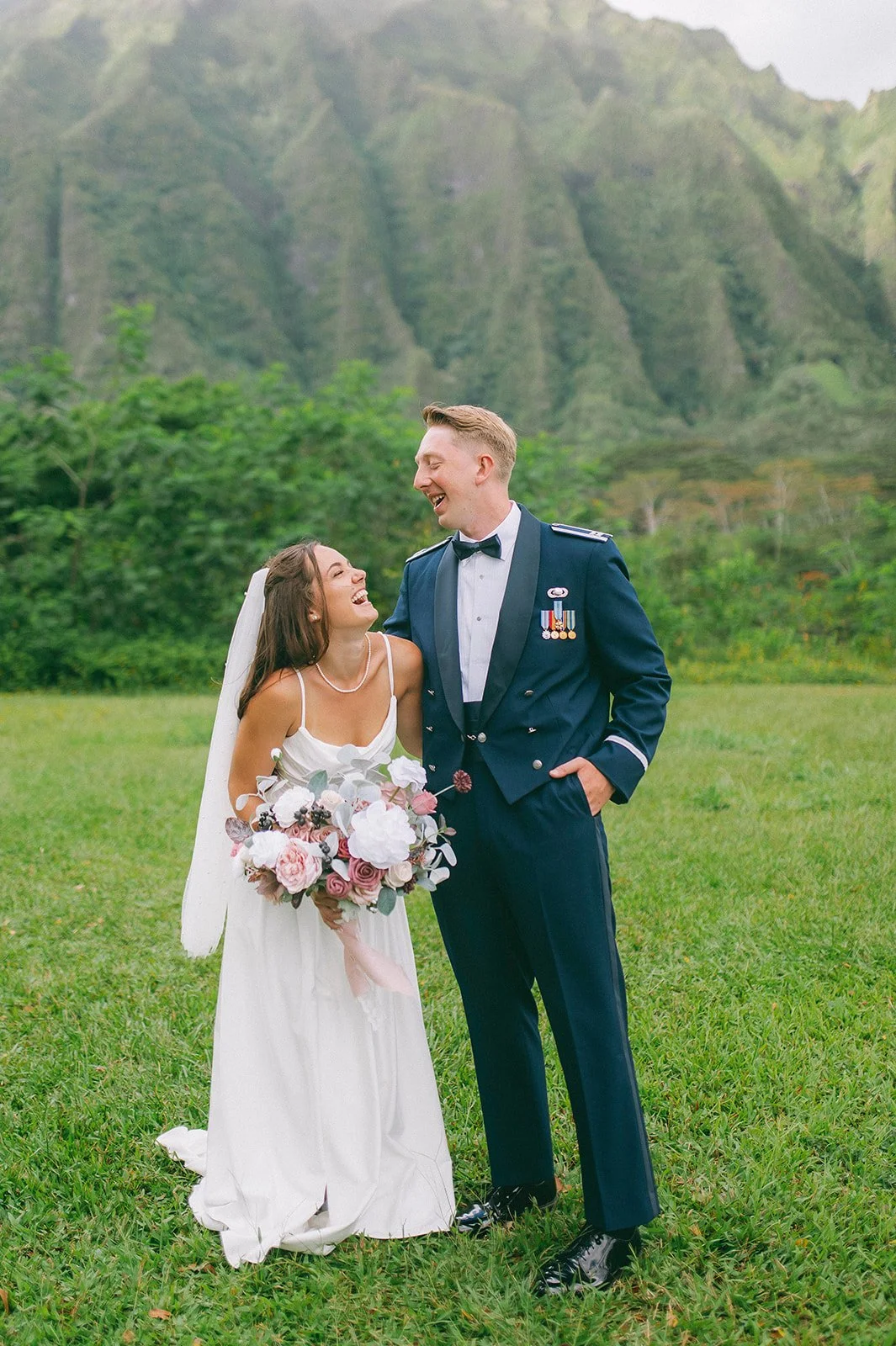 Bride and groom smiling together in a green outdoor landscape, with the groom wearing a formal military uniform.