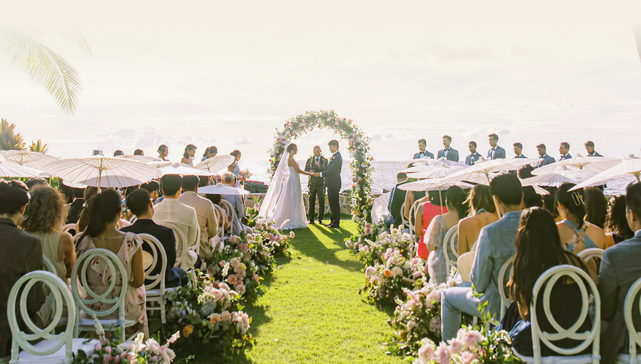 Outdoor oceanfront wedding ceremony with guests seated on both sides as the couple exchanges vows under a floral arch.