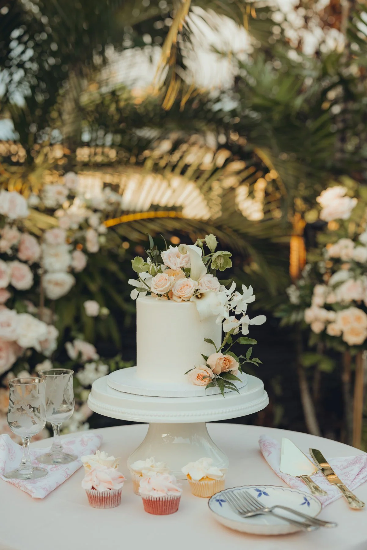 White wedding cake and floral cupcakes displayed among soft pink and white flowers.