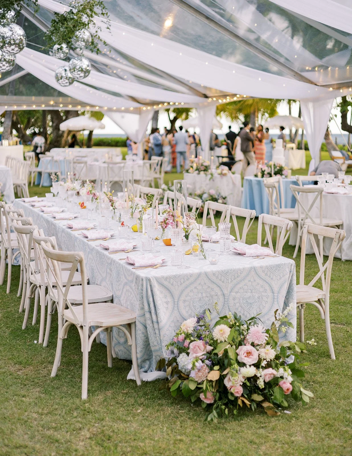 Elegant outdoor wedding reception table with floral arrangements, white chairs, and draped tent decor.