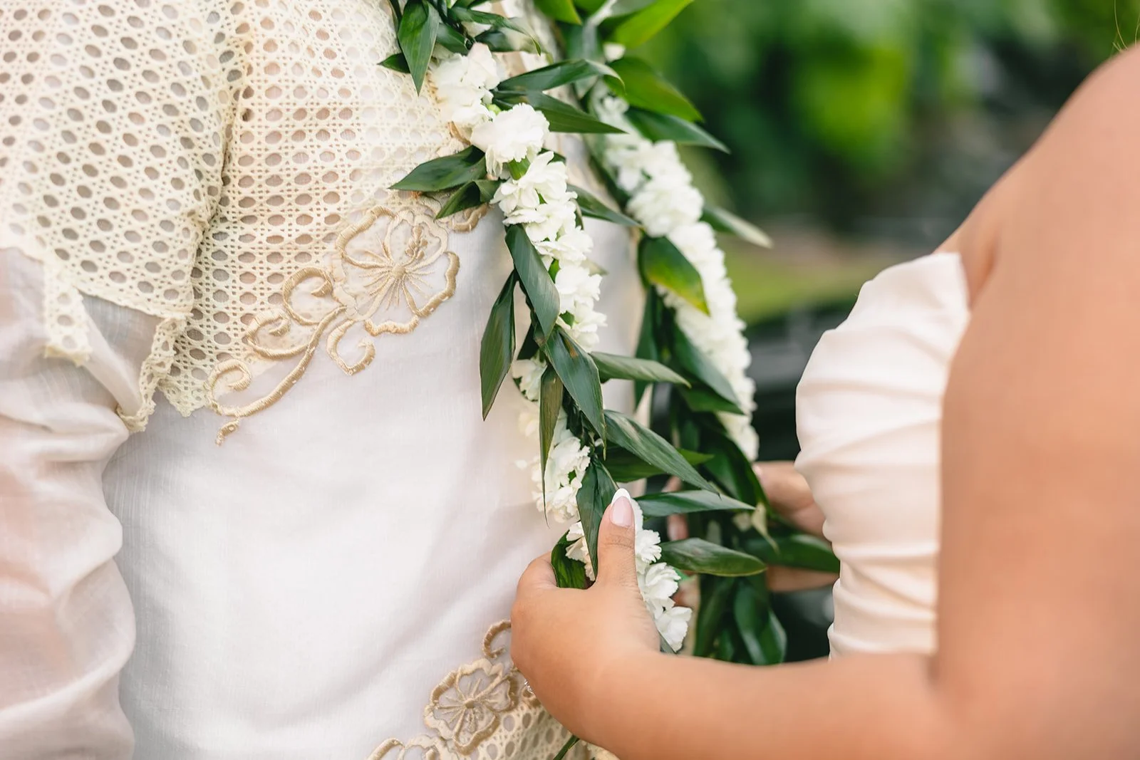 Close-up of a white wedding dress with green floral garland and delicate embroidered details.