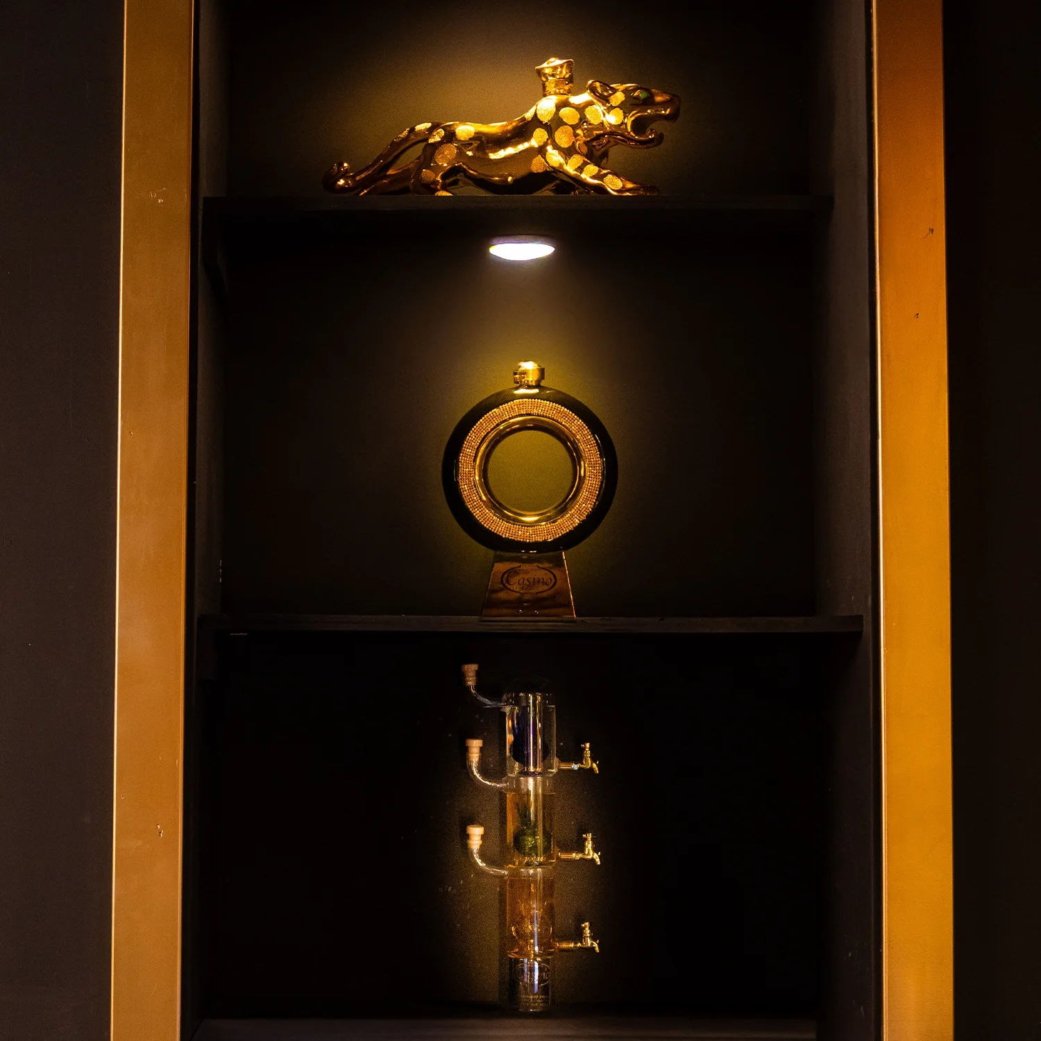 Decorative gold sculpture of a panther at the top, a gold and black round award or decorative object in the middle, and a glass apparatus with tubes and faucets at the bottom, all displayed in a dark cabinet with lighting highlighting each piece.