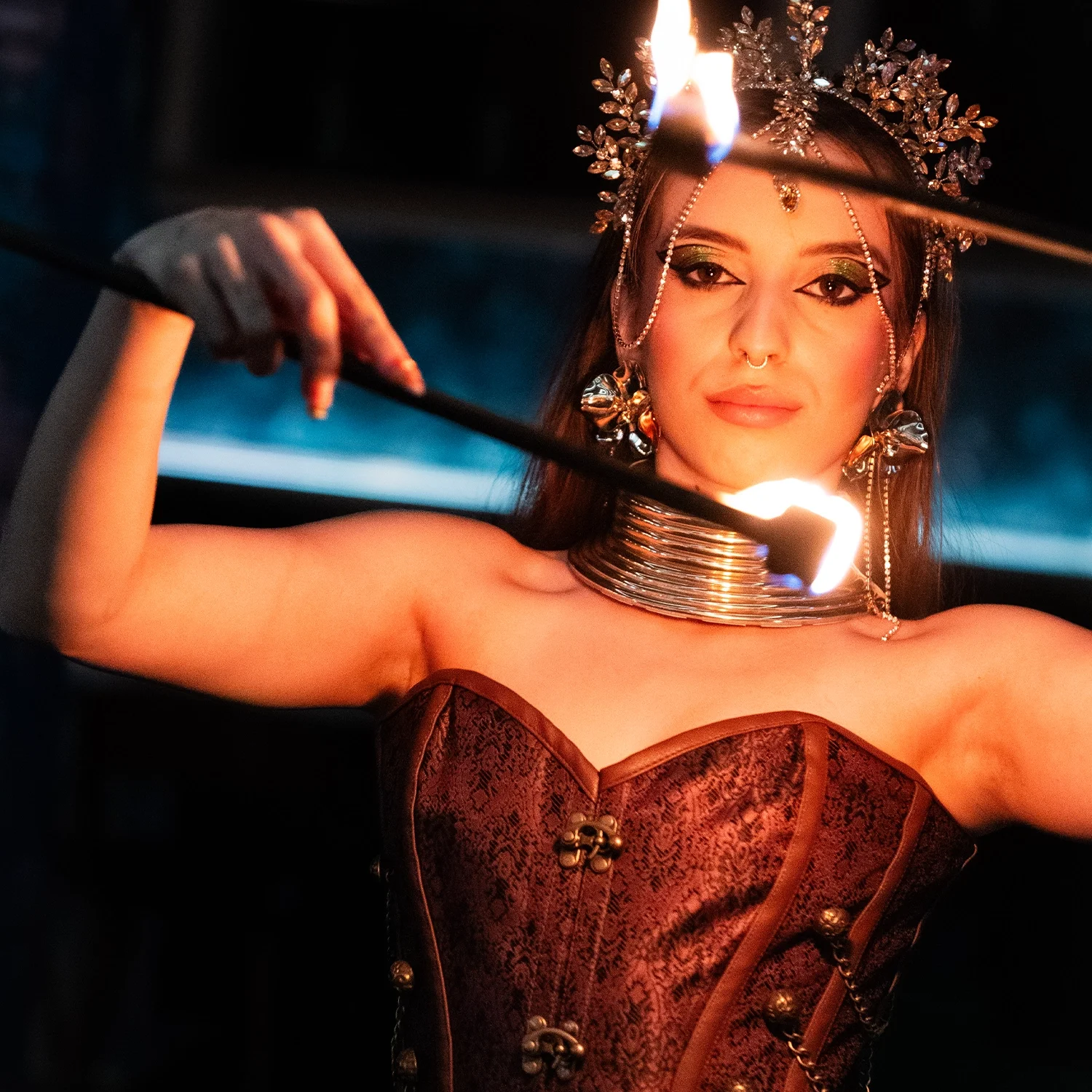 A woman dressed in an ornate, gothic-style outfit with metallic jewelry and a dark, floral headpiece, holding a stick with flames in a dark environment.