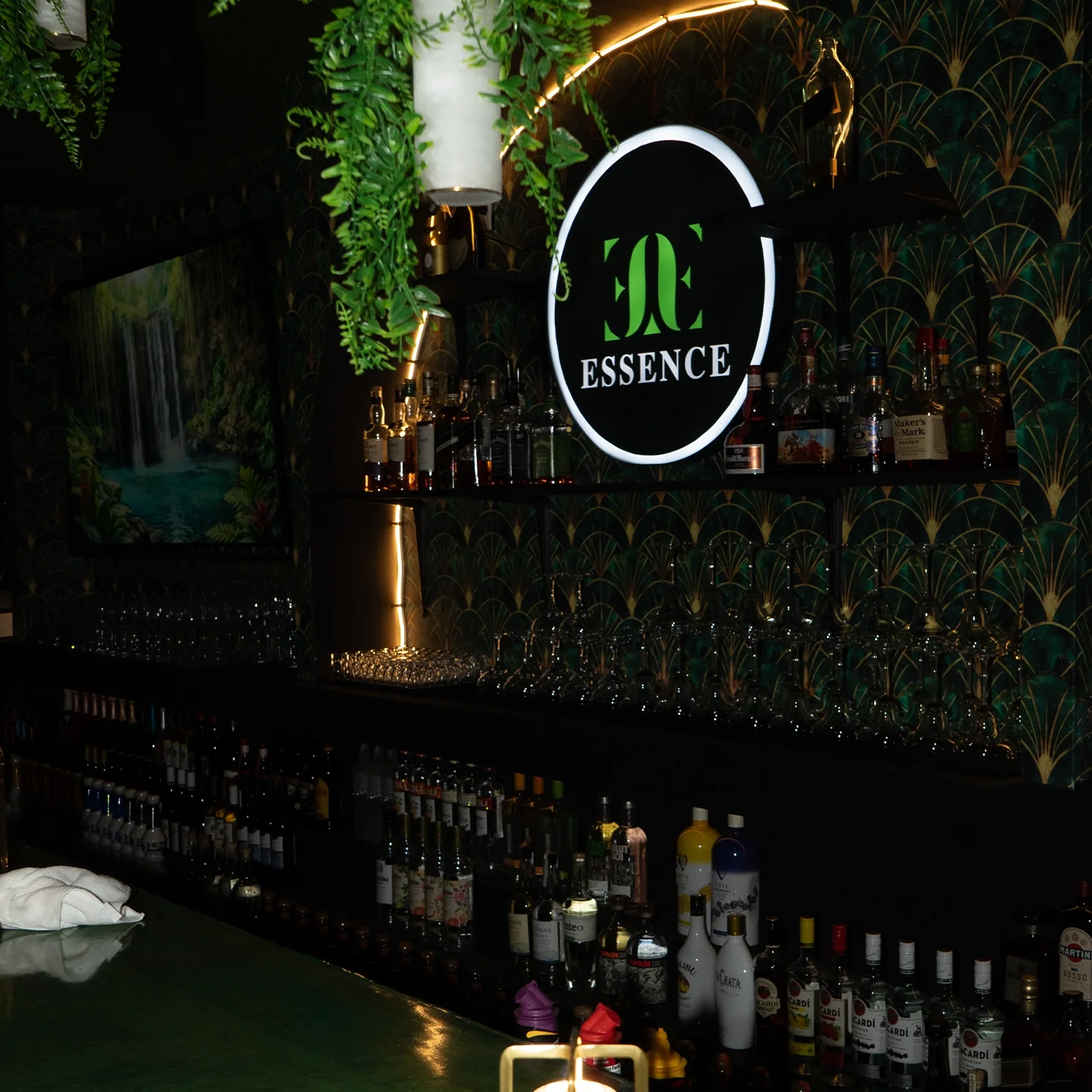 A bar with shelves of various liquor bottles, glassware, and a illuminated sign that reads 'ESSENCE' in a dark, decorated setting.