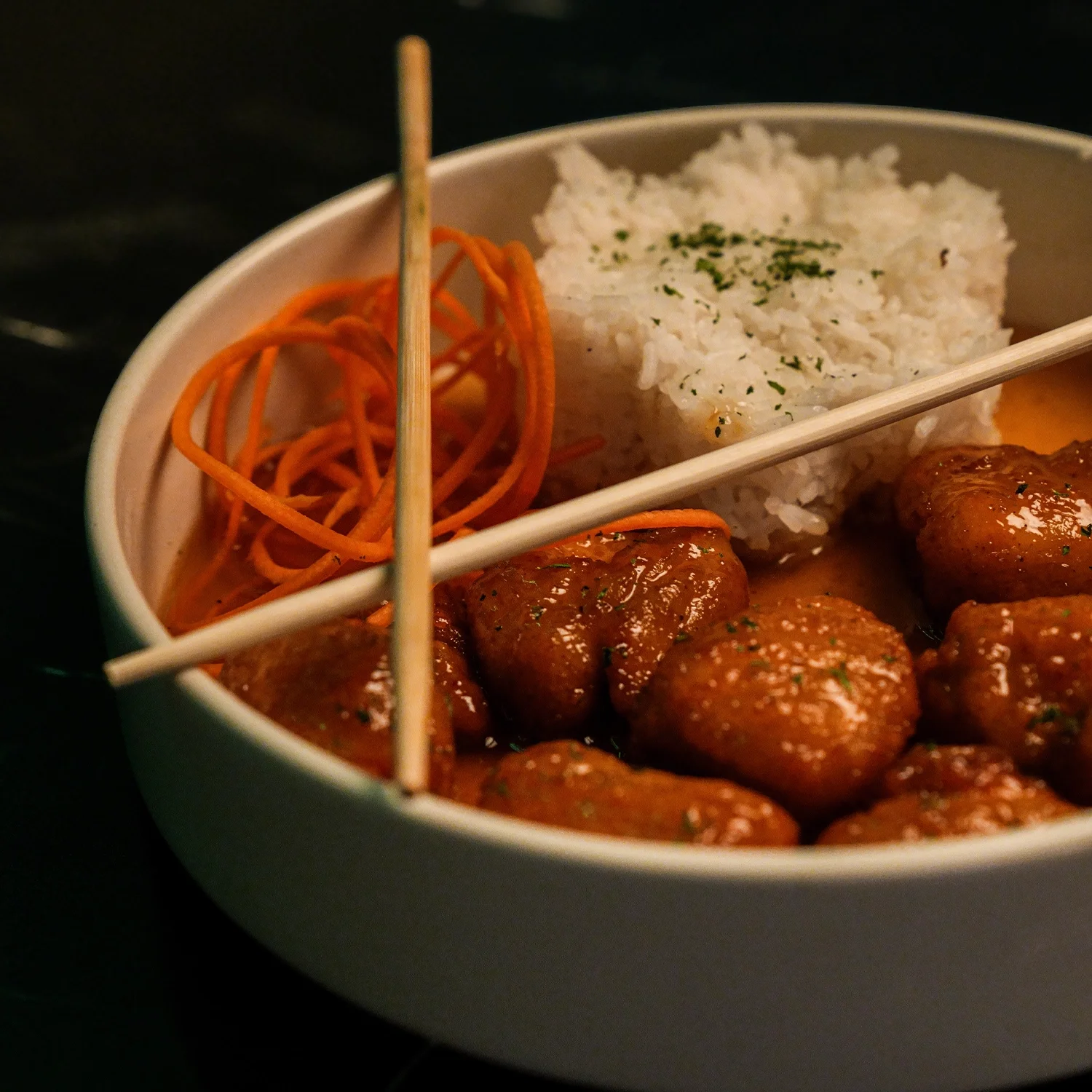 Asian-inspired meal in a white bowl with rice, orange noodles, and glazed meatballs, edited with a dark background.
