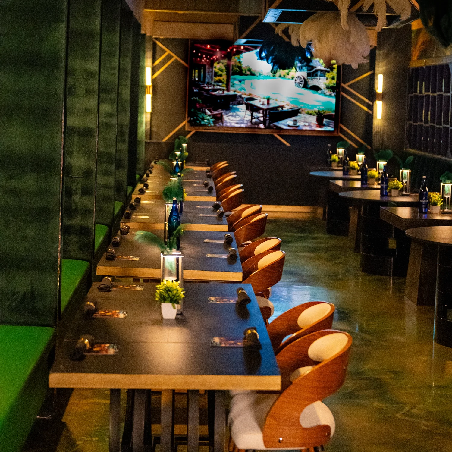 Empty restaurant with a row of tables set for dining, small lamps, plants, and a large screen displaying an outdoor scene.