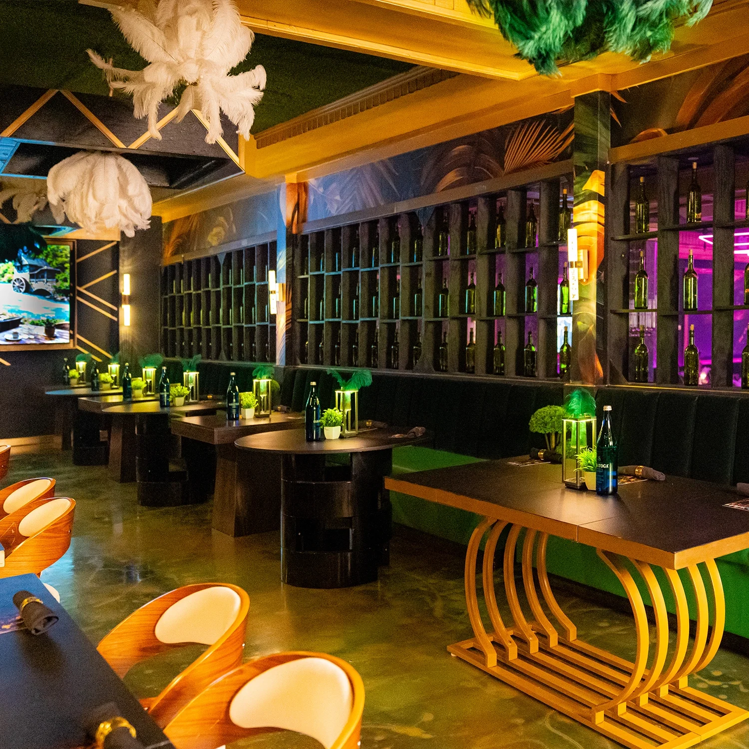 Interior of a restaurant or bar with dim lighting, decorated with green plants and bottles on shelves, with round tables set with water bottles, small plants, and lanterns, and wooden chairs with white cushions.