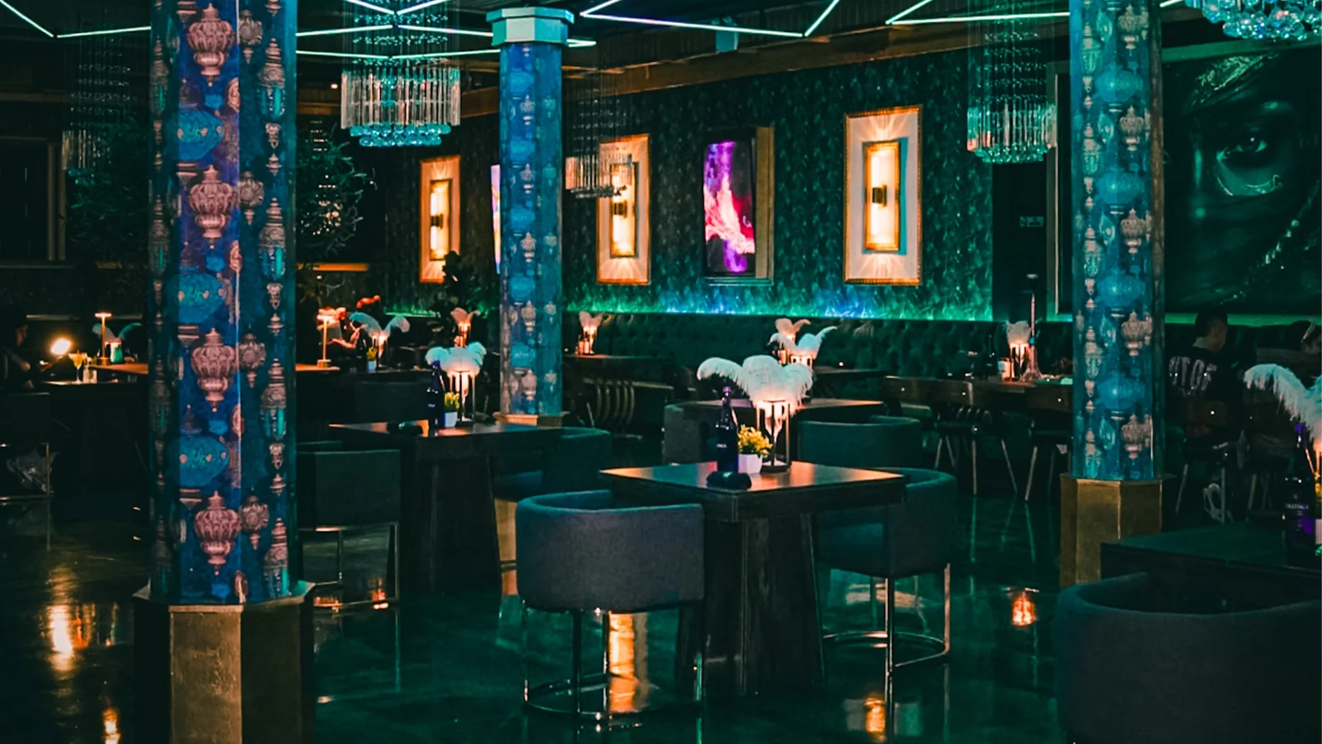 Interior of a dimly lit upscale restaurant or lounge with dark green walls, illuminated picture frames, plush green seating, and tables decorated with white feather centerpieces, bottles, and flowers.