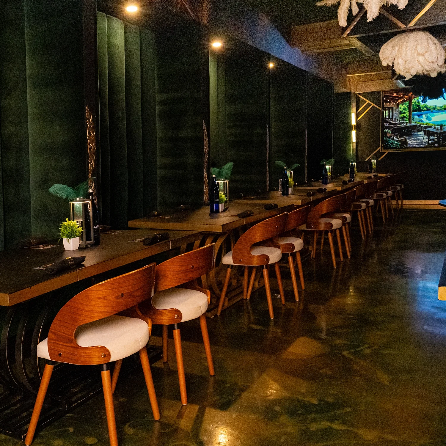 An interior view of a restaurant with a long wooden counter and matching bar stools with beige cushions. The counter is decorated with small potted plants, bottles, and table settings. The wall behind the counter has green panels, and there is a larg
