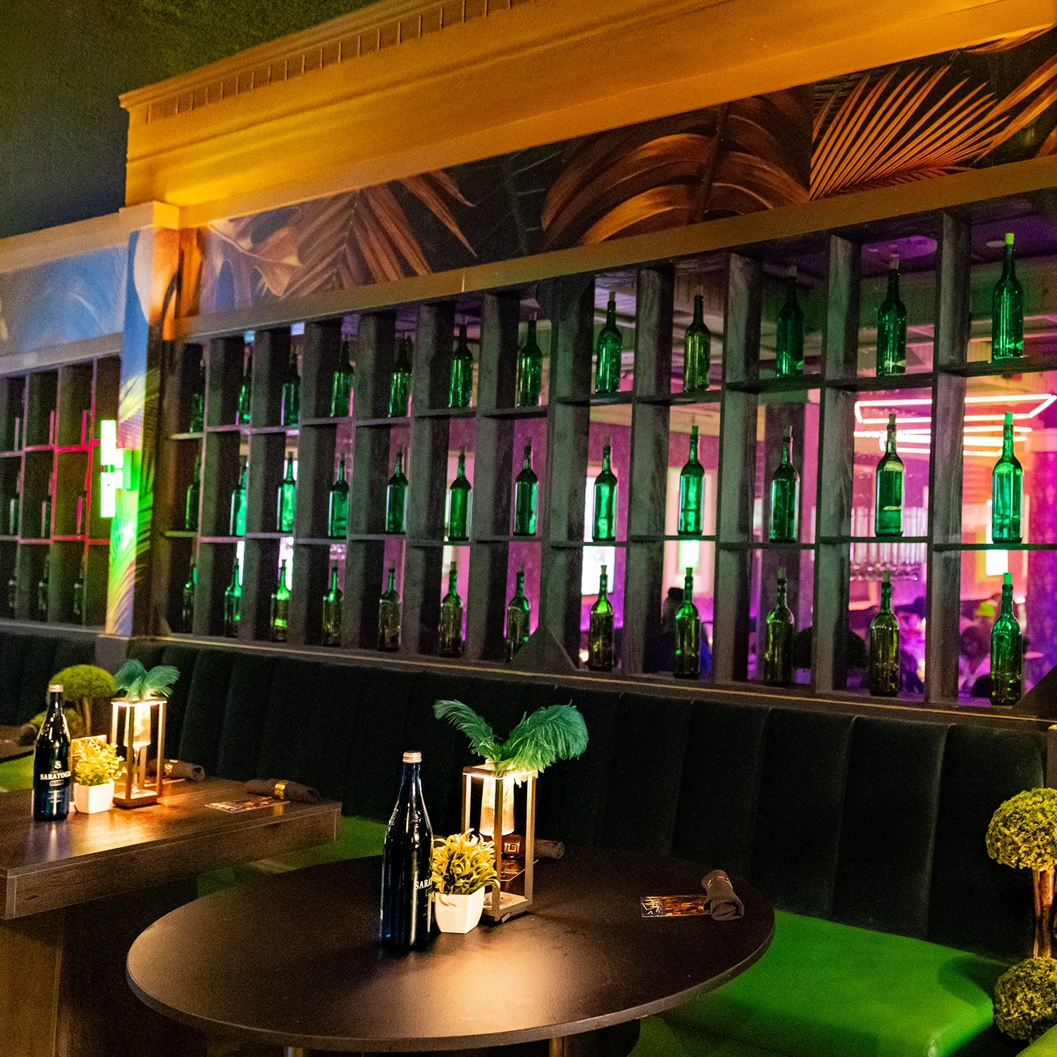 Interior of a bar or restaurant with a decorative wall featuring rows of green bottles, illuminated with colorful purple and pink neon lights, and tables decorated with plants and bottles.
