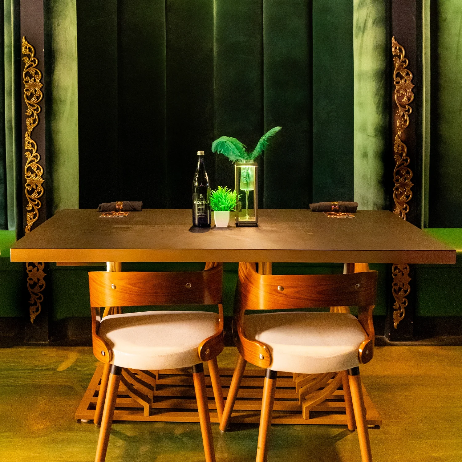 A dining table with two chairs, decorated with a bottle, a potted plant, and a decorative lamp, set against a green and black wall with gold ornate accents.