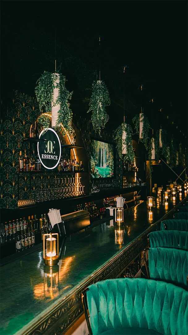 A dimly lit bar with green velvet chairs, hanging plants, and a backlit sign reading 'ESSENCE' above bottles of liquor.
