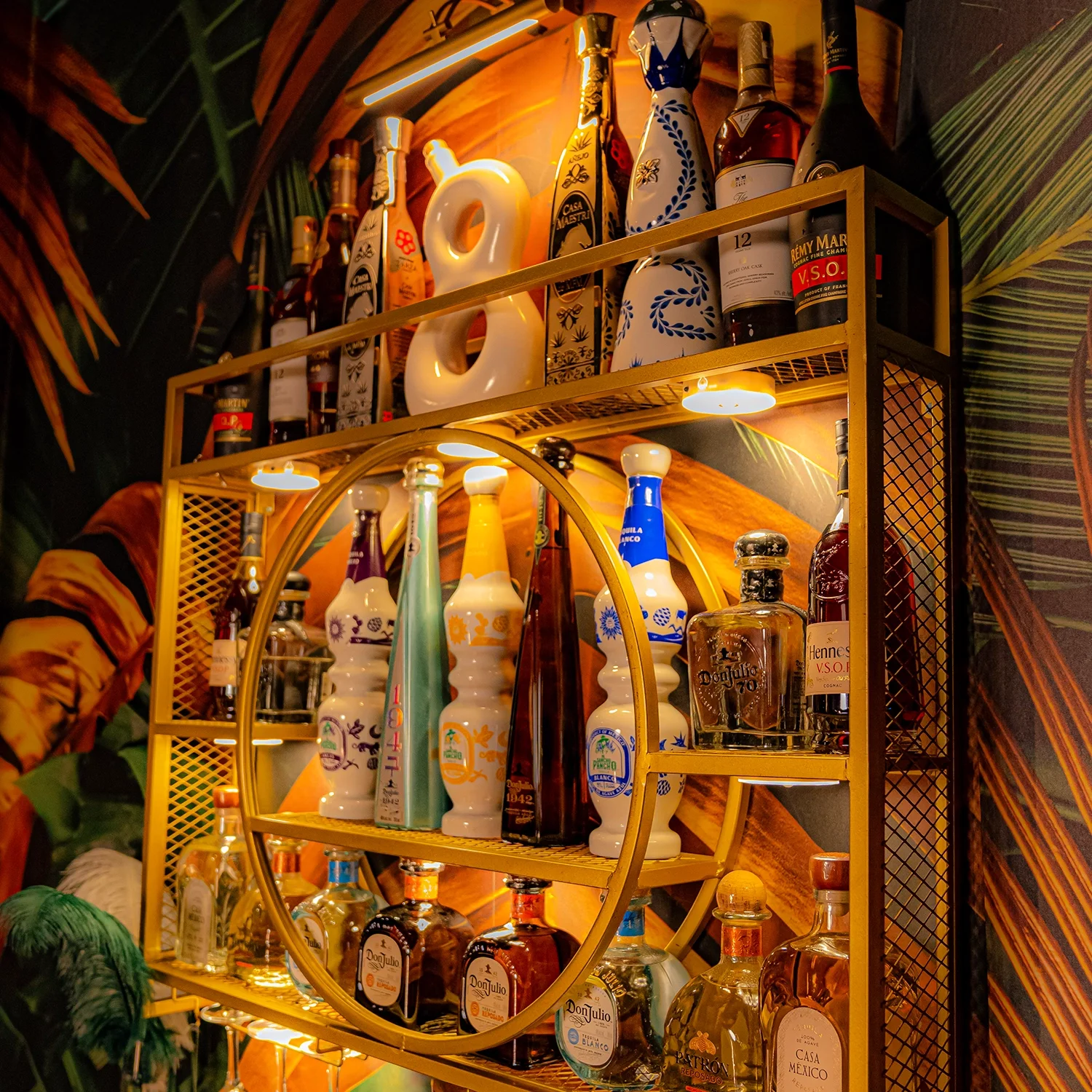 A gold shelving unit with liquor bottles and decorative vases, illuminated with warm lighting, against a colorful tropical-themed wall.