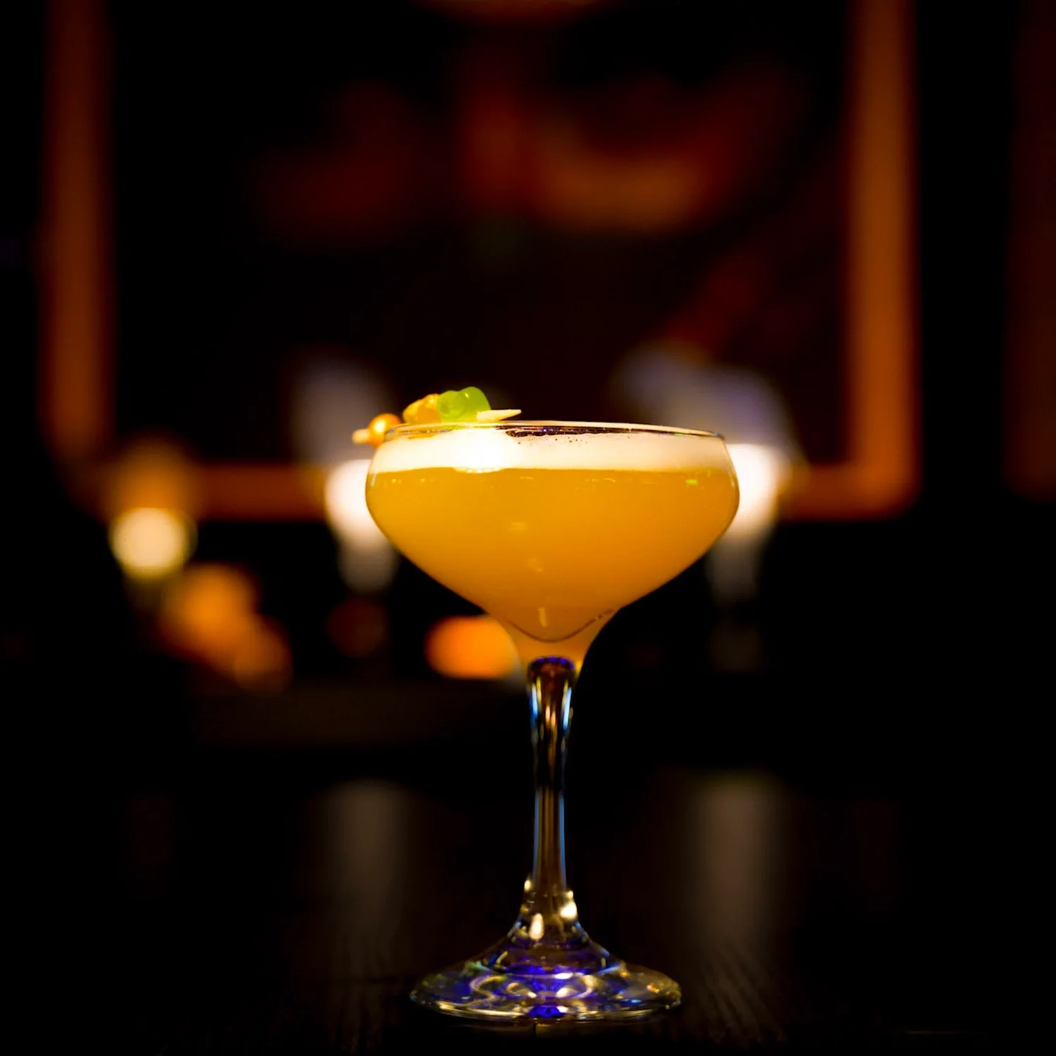 A yellow cocktail in a coupe glass garnished with a small green fruit and a cherry, set against a dark, blurred background.