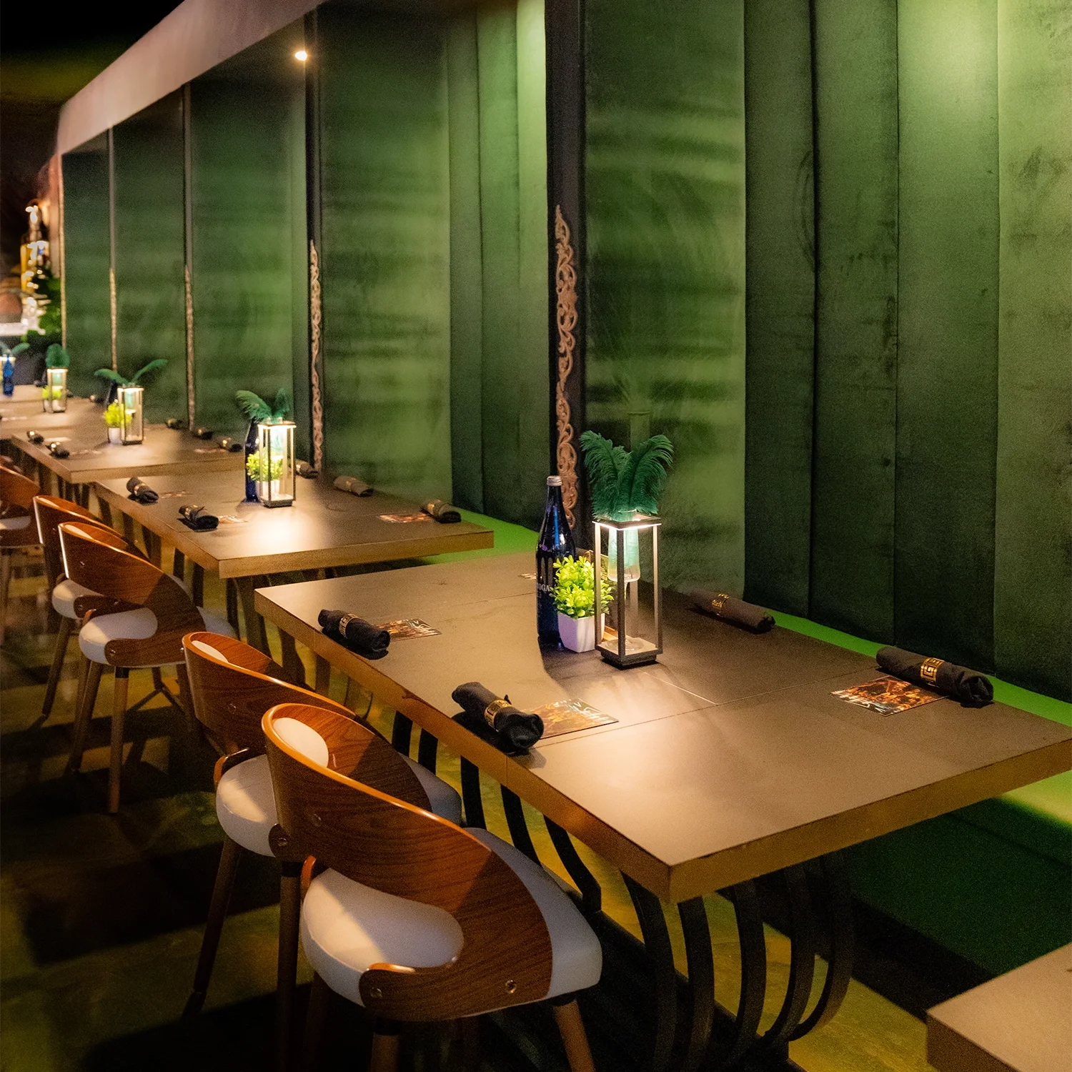 Empty restaurant table set with black napkins, lanterns, bottles, and decorative plants against a green wall.