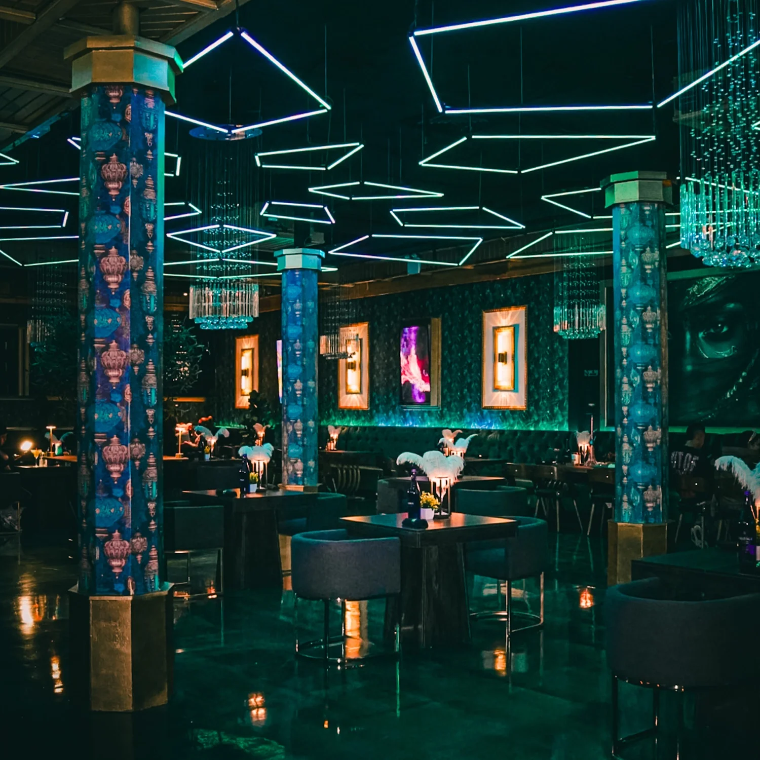 Interior of a dimly lit upscale lounge or bar with neon geometric ceiling lights, decorative columns, framed artwork on the walls, plush seating, and small tables with flowers and drinks.