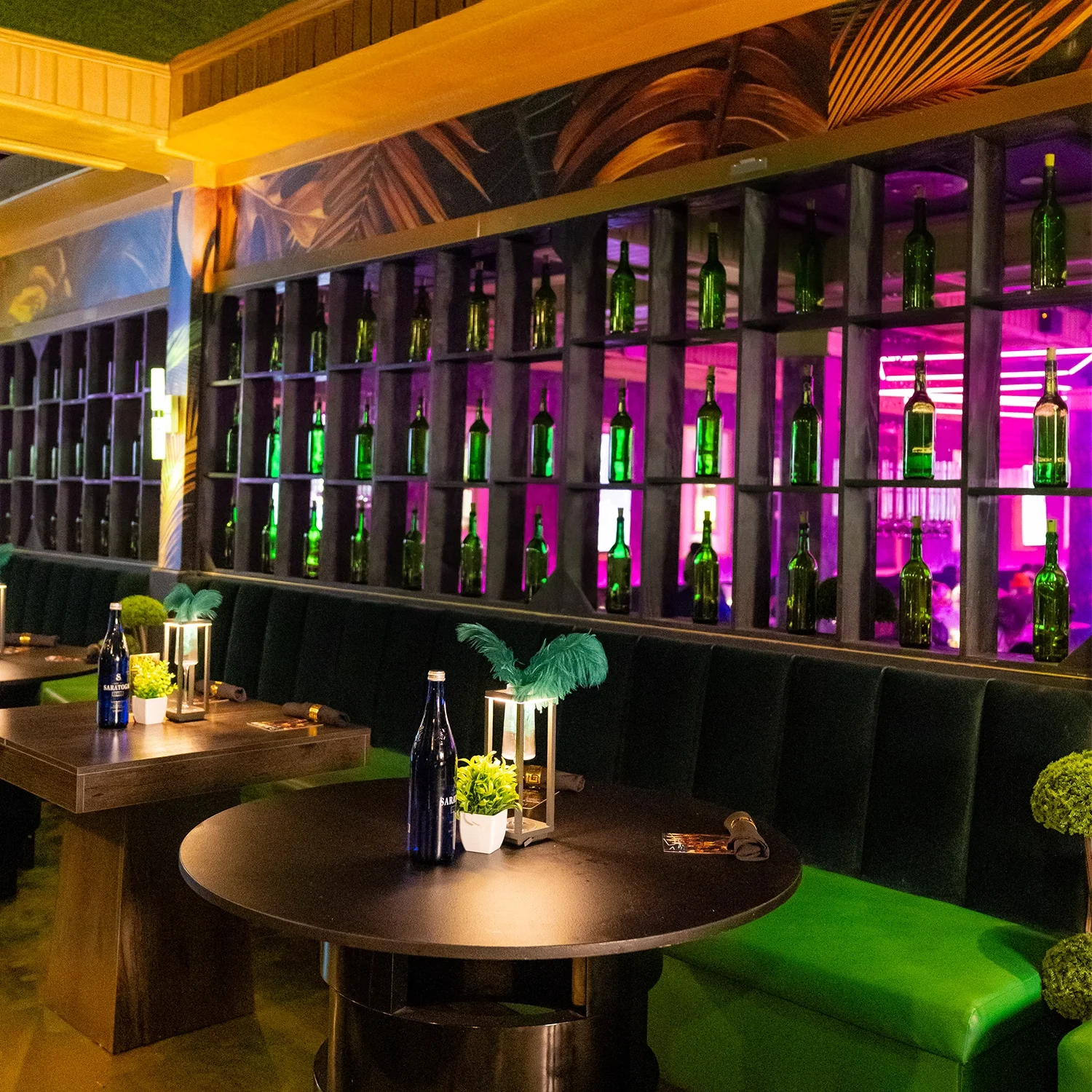 Nighttime view of a restaurant interior with tables, green upholstered seating, small potted plants, and decorative lamps with feathers, with a wall display of green bottles and neon pink lights in the background.