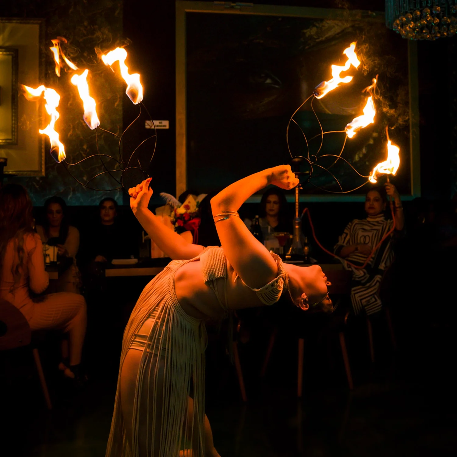 A female fire performer flipping flaming fans in a dimly lit room, with an audience seated around her.