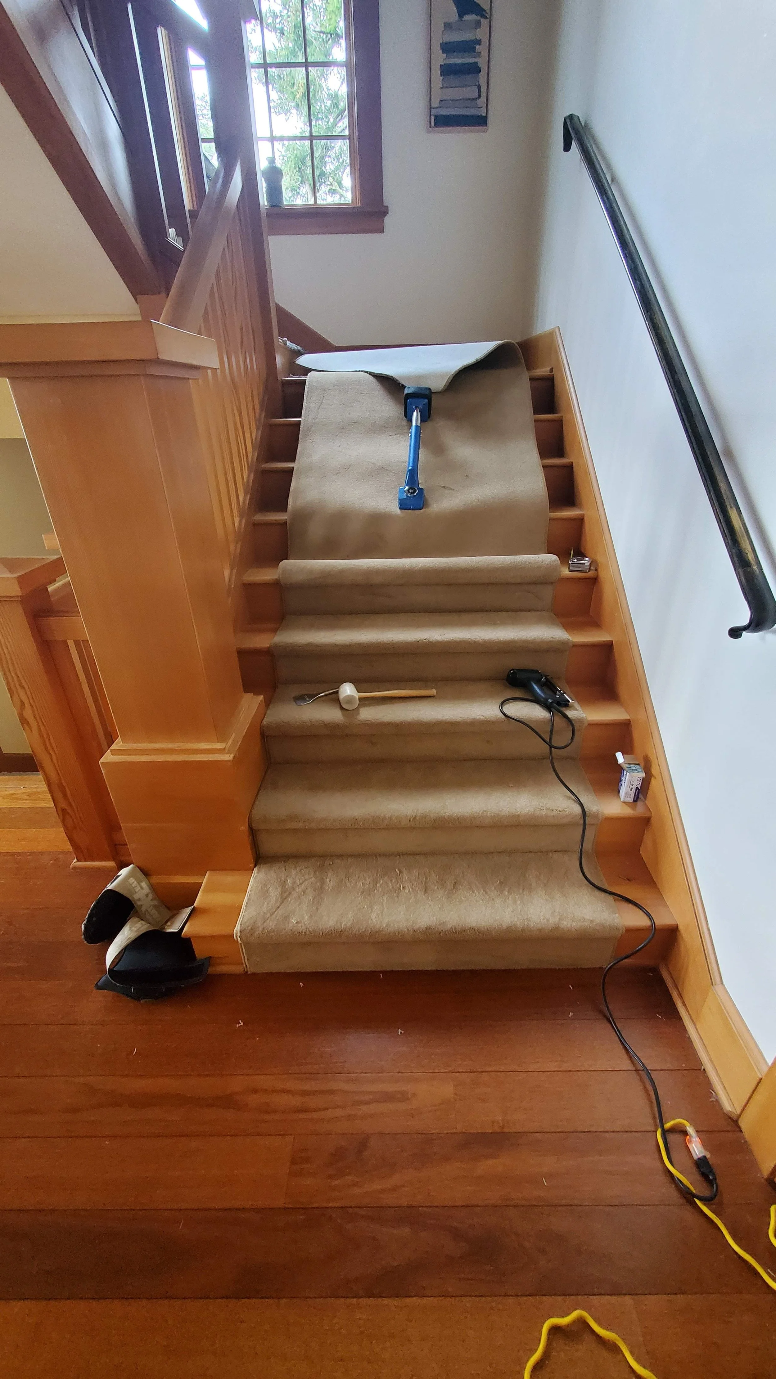 The image shows a staircase with brown carpeting, some tools, and an extension cord on the steps. To the left, part of a wooden railing and a window giving natural light are visible.