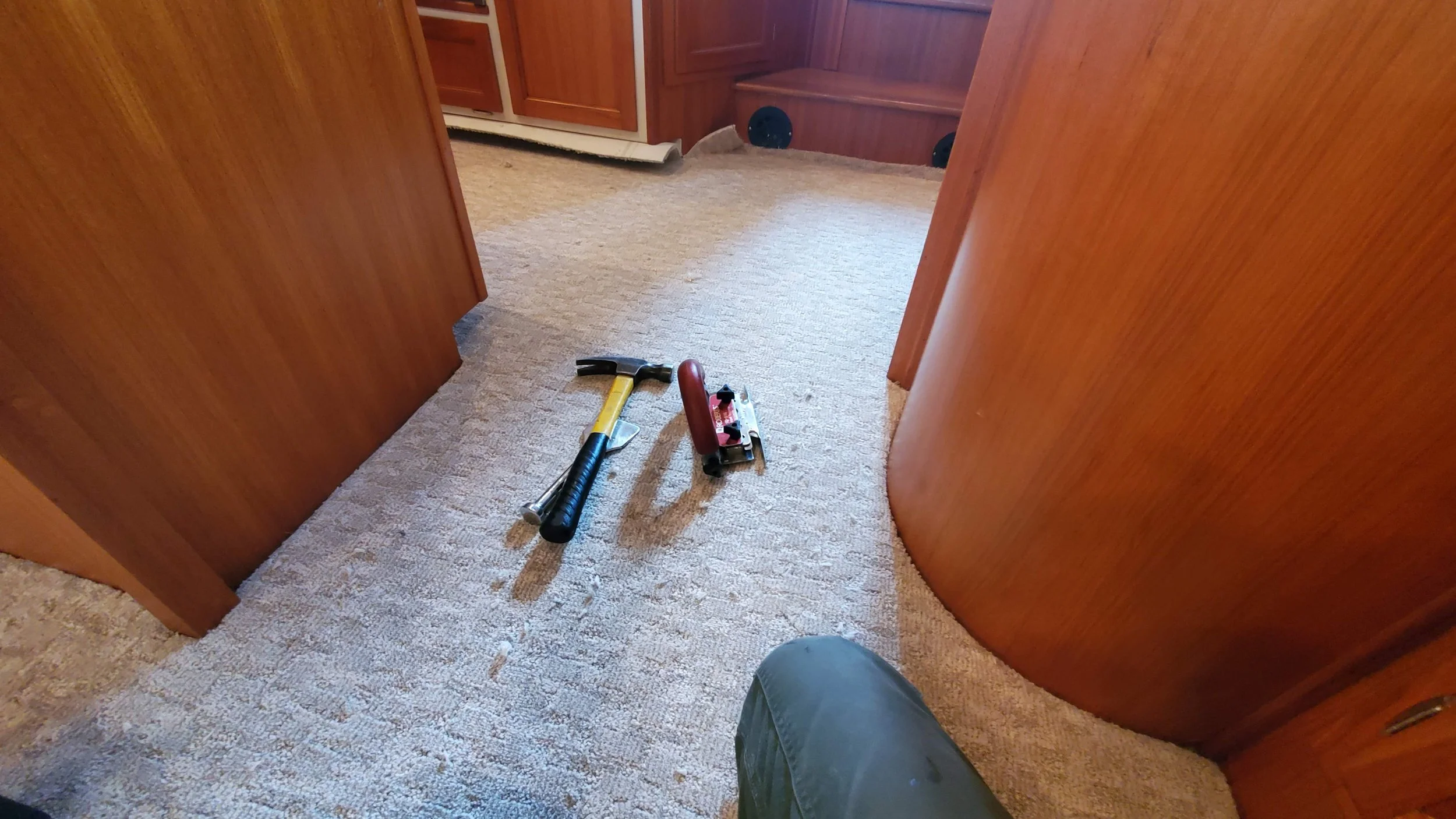 Interior view of a room with wooden walls, carpeted floor, and tools including a hammer and a tool with a red handle on the floor.