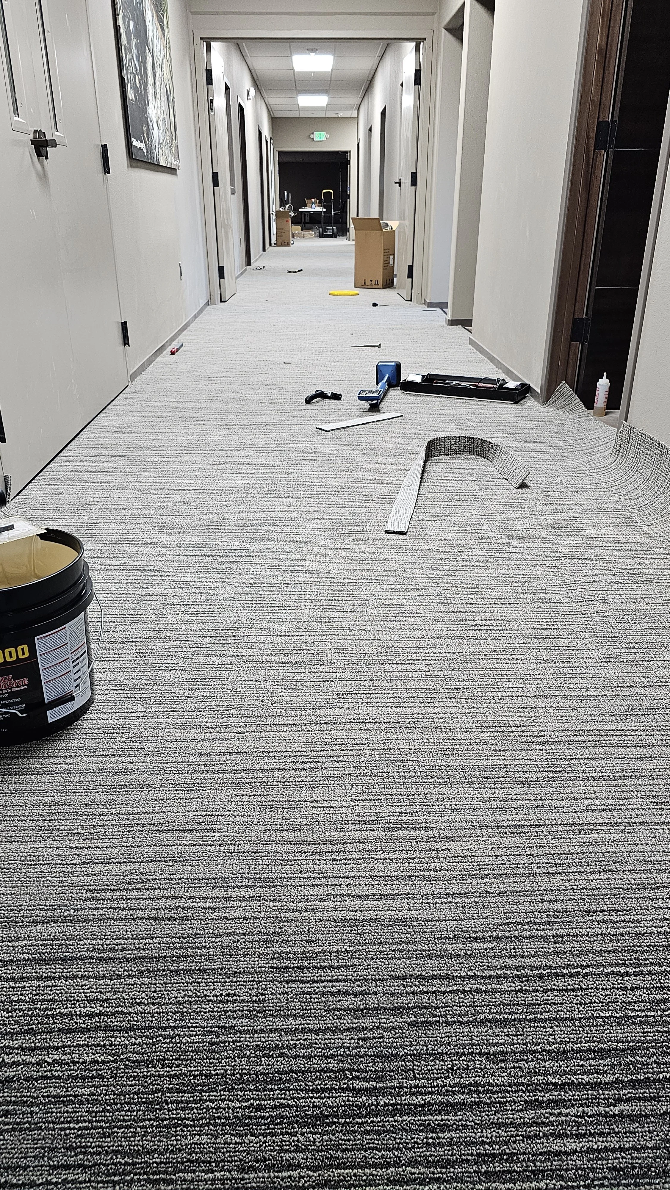 A hallway with scattered tools and construction materials, including a black bucket, a measuring tape, a box, and various tools on the floor, leading to a room with a sink and boxes.