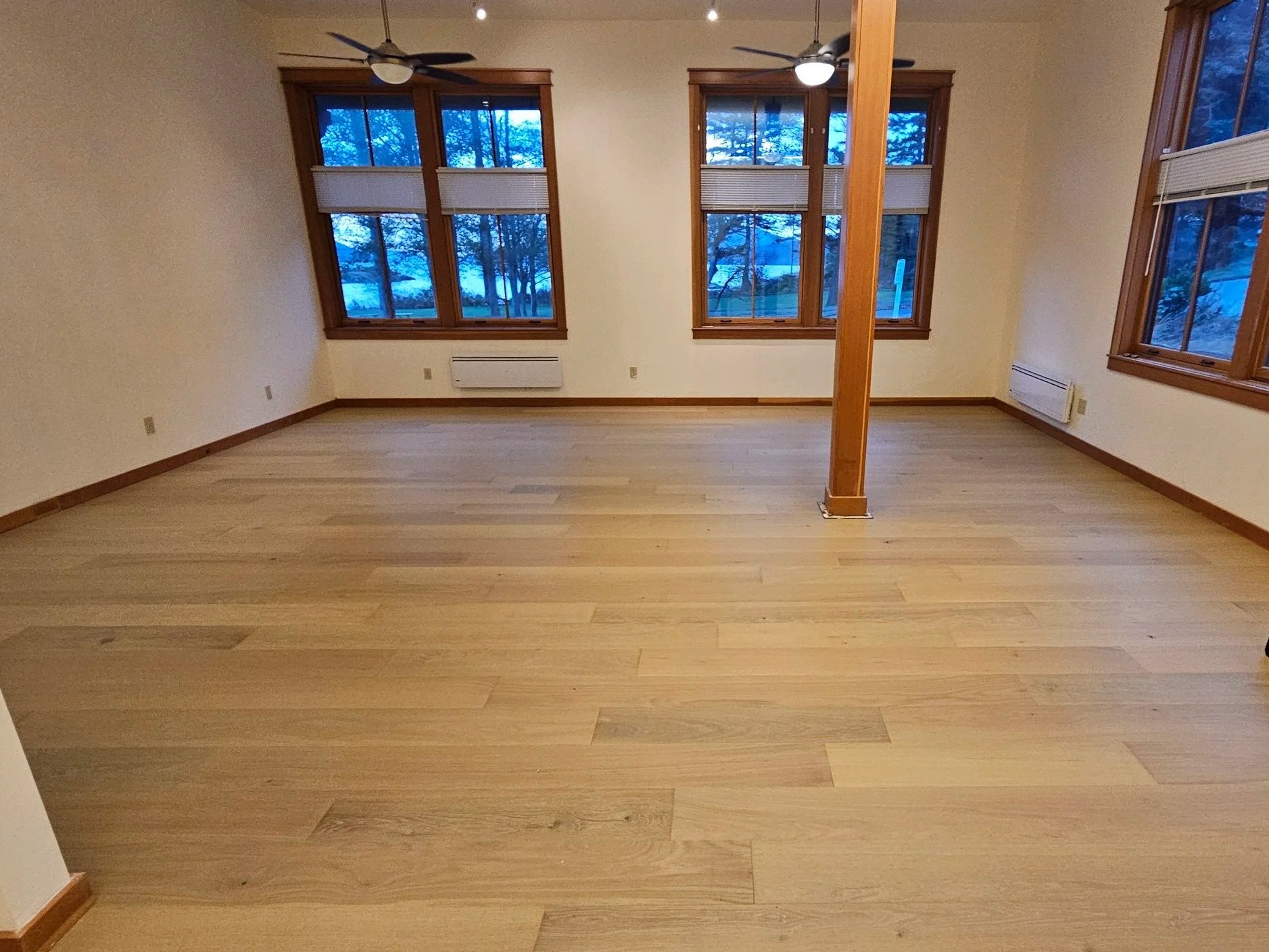 An empty room with wooden floors, three large windows with wooden frames, and a wooden support beam in the center. Two ceiling fans are attached to the ceiling, and an air conditioner is visible beneath the windows.