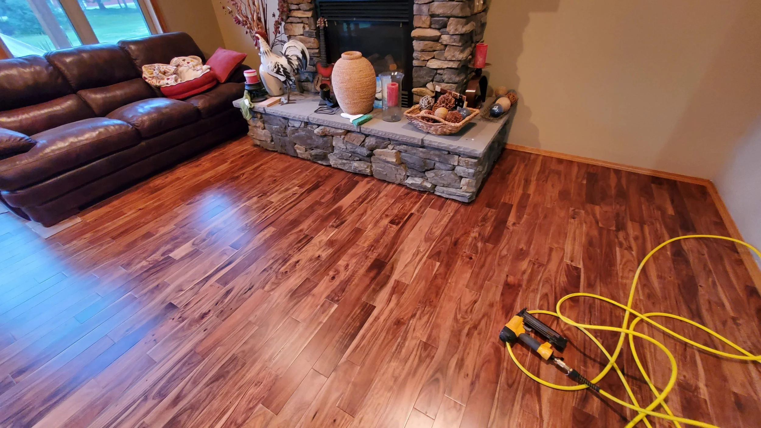 The image shows a living room with polished hardwood floors, a brown leather sofa with pillows and blankets, and a stone fireplace with decorations including vases and candles. There is a yellow extension cord and a power tool on the floor.