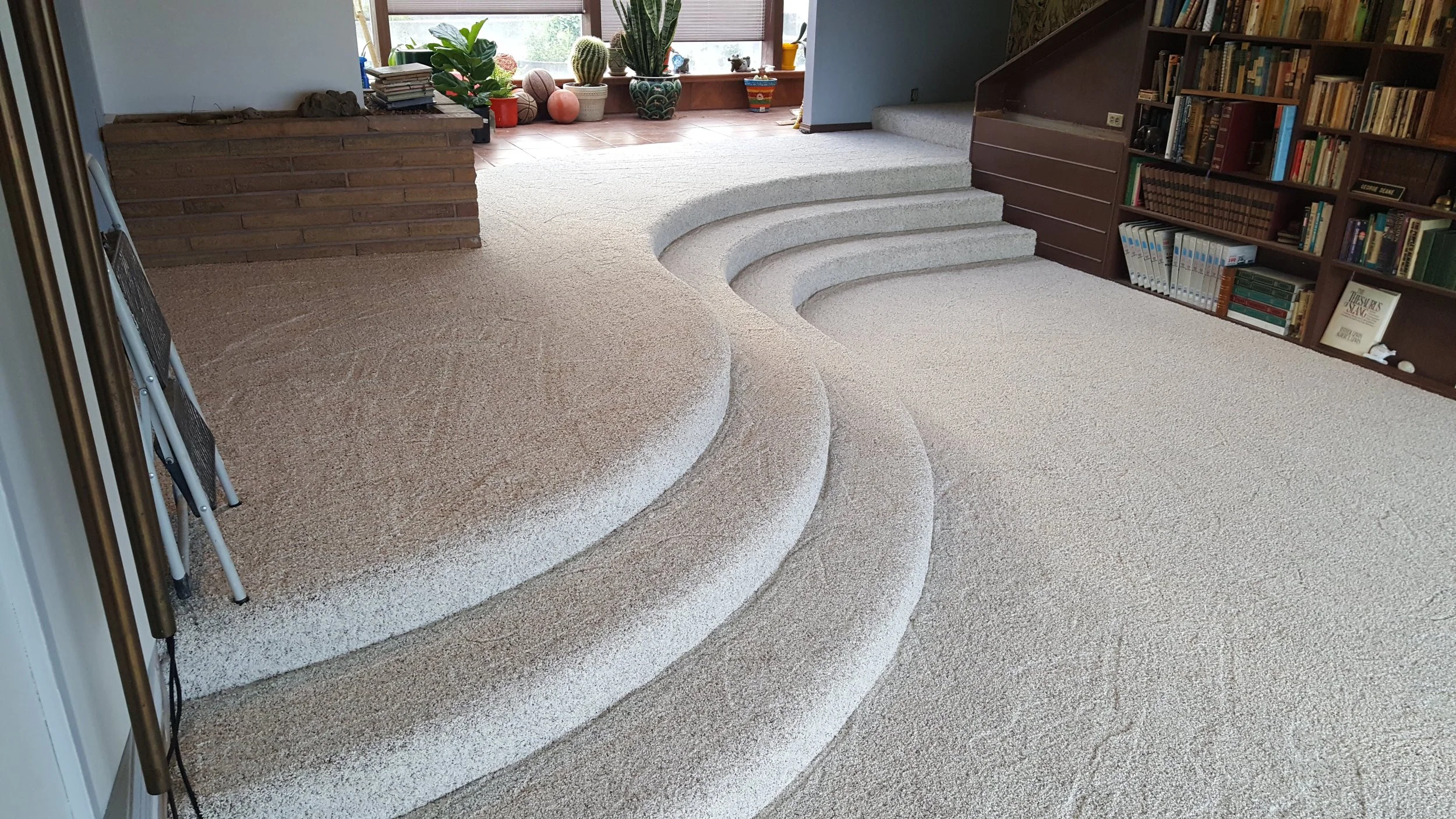 Room with curved carpeted steps, a brick platform, a bookshelf filled with books, a window with plants and decorative objects, and a small staircase leading to another area.