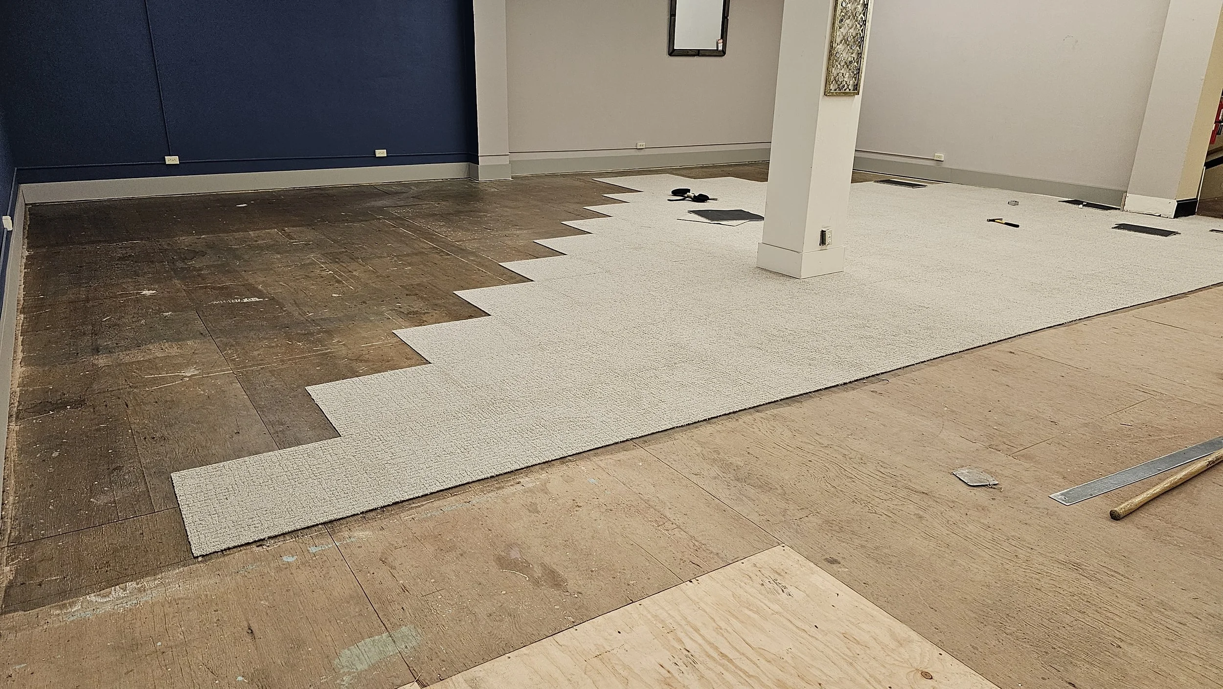Room under renovation with partially installed white carpet over wooden flooring, tools and materials scattered around.