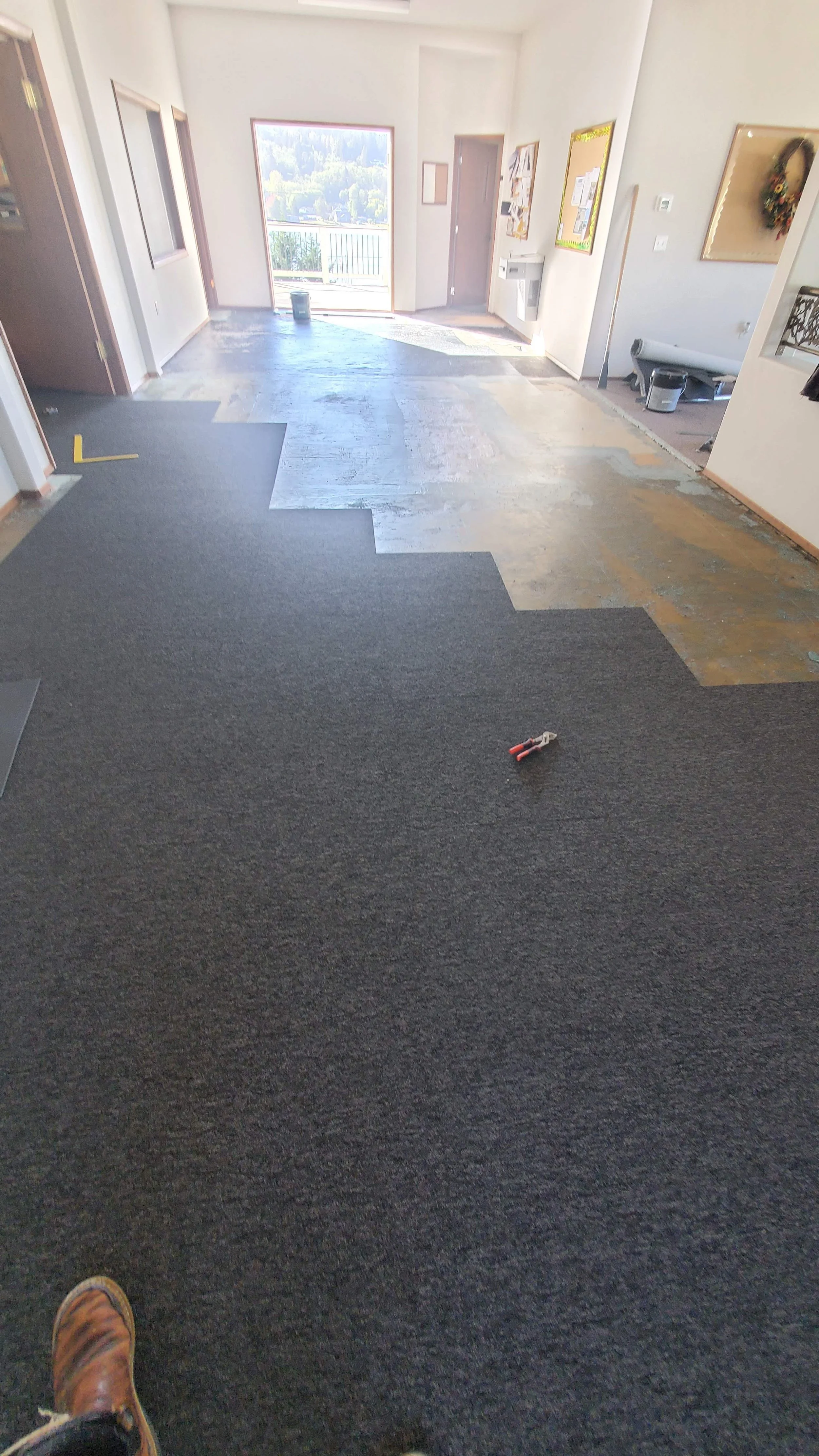 This image shows the interior of a building with a partially renovated floor. The room has white walls, and there is a large window or door at the front letting in sunlight. The floor is partially covered with dark gray carpeting, while the rest is in subfloor or underlayment, indicating ongoing flooring work. Construction tools and materials are visible on the floor, and the room appears to be in the process of flooring installation or renovation.