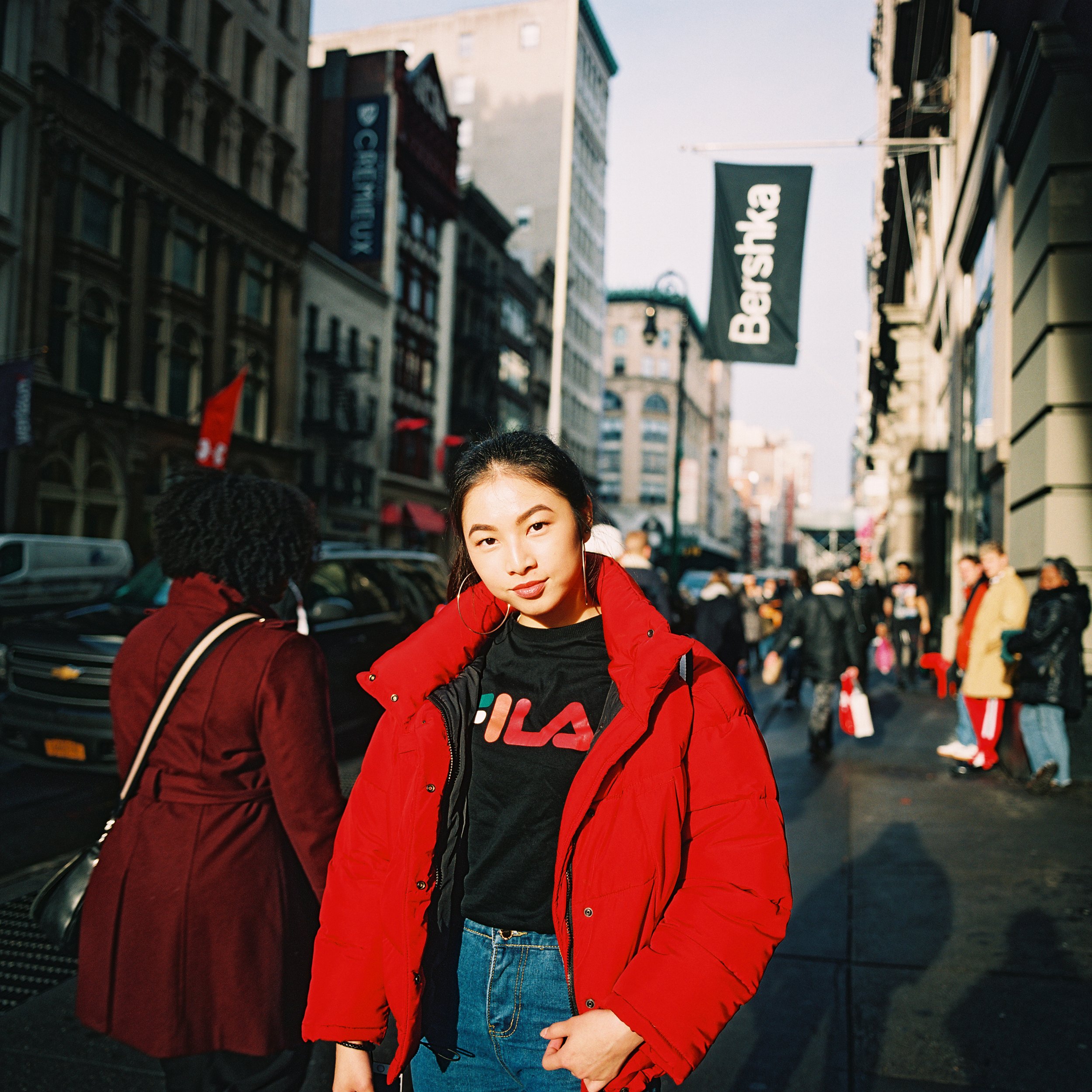 Jovencita en una calle de ciudad con edificios altos, vestida con chaqueta roja y camiseta negra con logotipo, rodeada de peatones y autos, con tienda 'Bershka' visible.