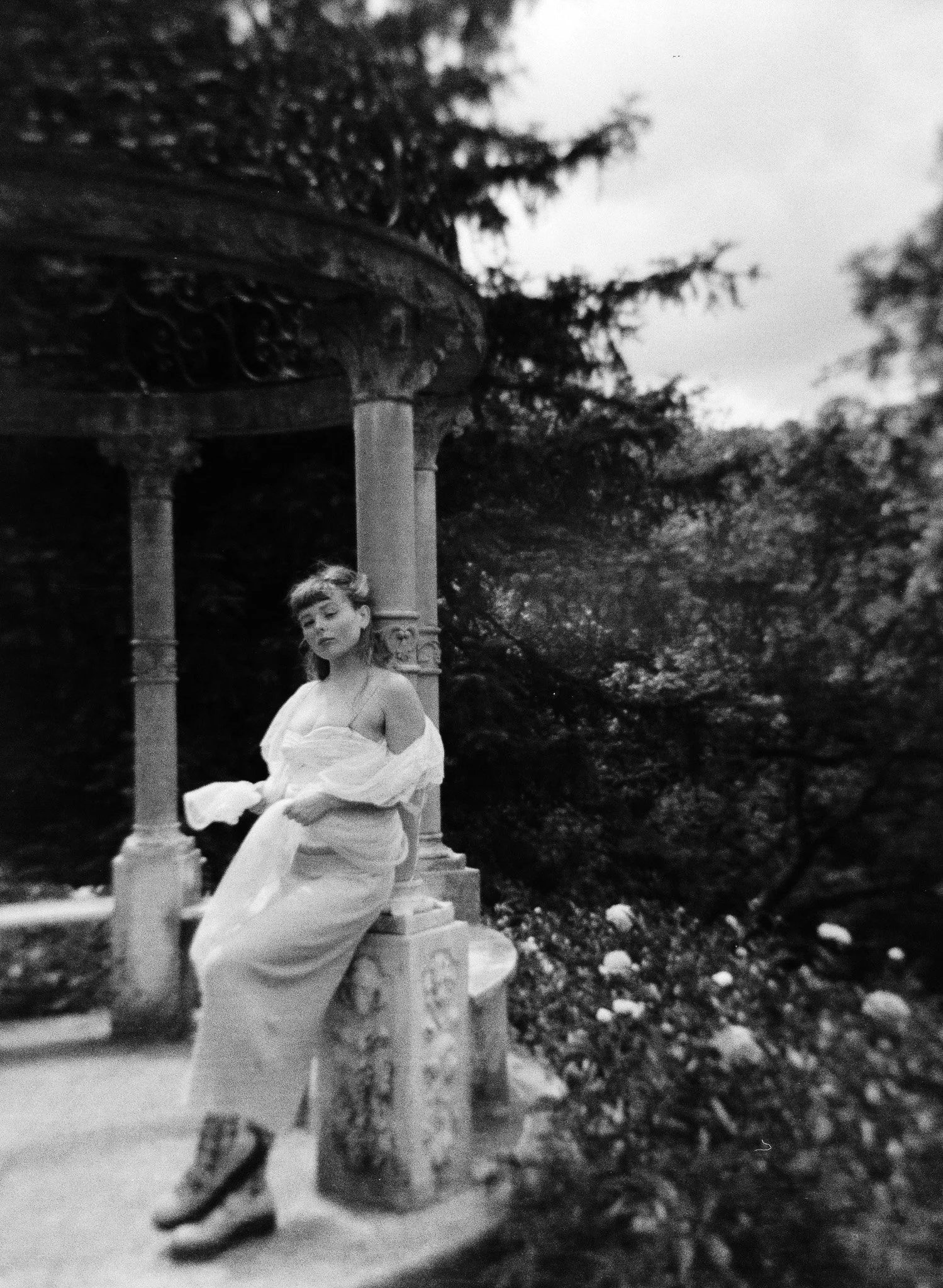 Una mujer joven con un vestido largo, sentado en un banco de piedra junto a una estructura de columnas en un entorno natural con árboles y flores a su alrededor.