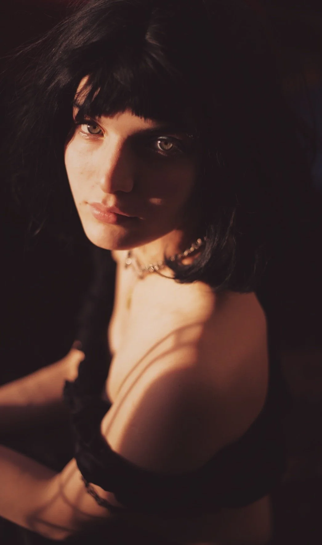 Retrato de una mujer con cabello negro y liso, con sombra y luz suave que resalta su rostro y hombro, vistiendo ropa oscura y con un collar plateado, en un ambiente oscuro.