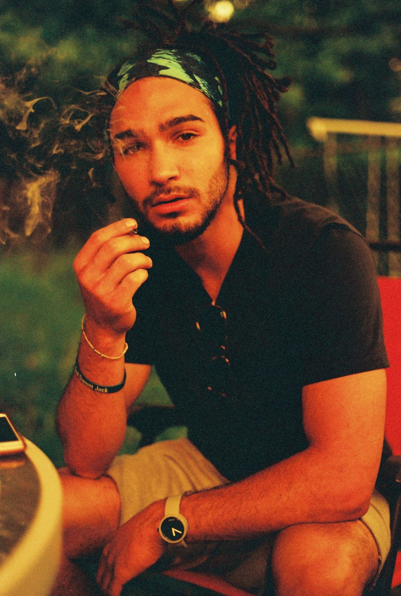 Joven con cabello rasta y bandana, sentado en una silla roja, sosteniendo un cigarrillo, con gafas colgadas en su camiseta y reloj blanco en la mano, en un entorno con iluminación cálida.