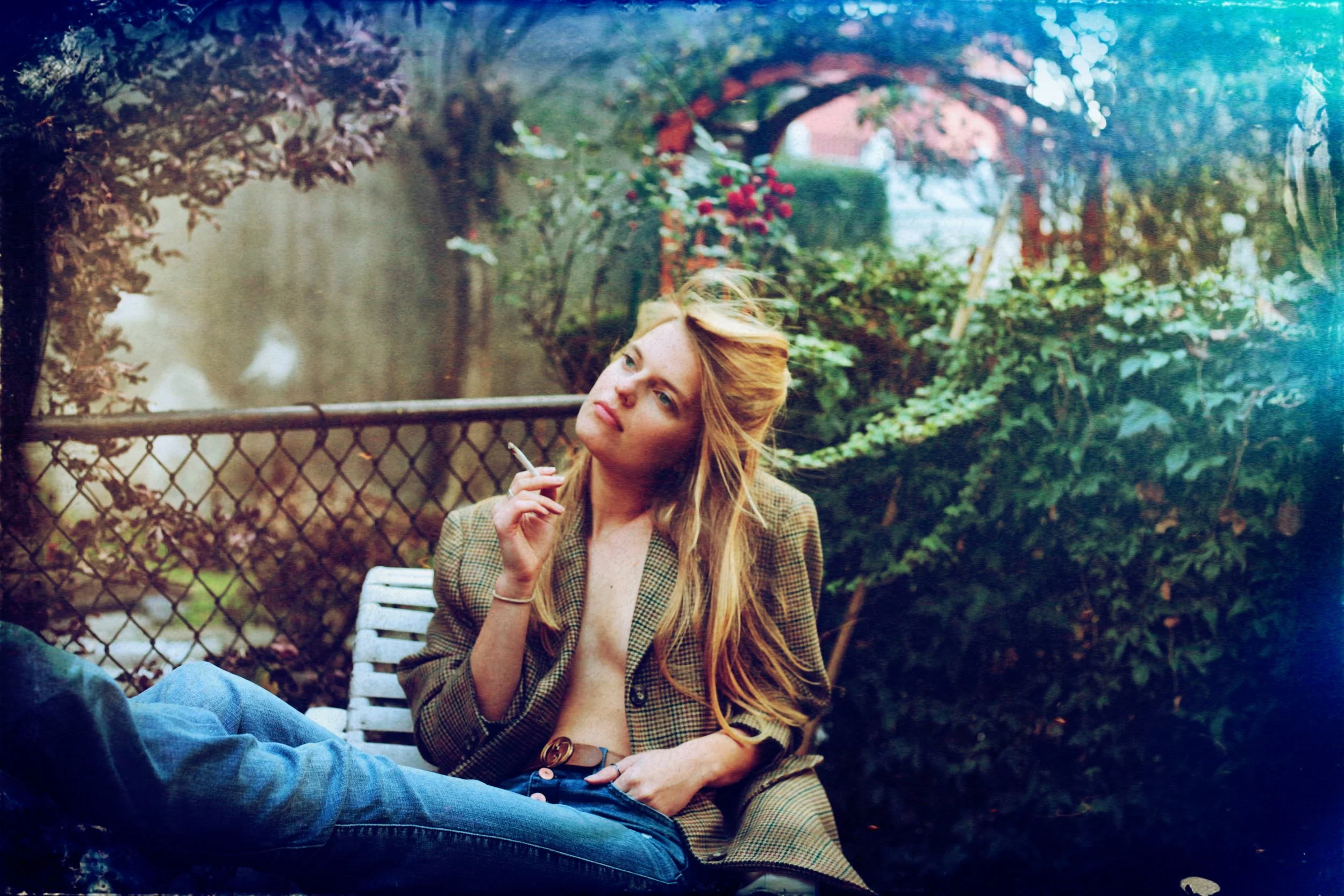 Una mujer con cabello rubio y largo, con una chaqueta a cuadros, se encuentra sentada en un banco de jardín, sosteniendo un cigarro y mirando pensativa hacia un lado. El fondo tiene plantas y un cerco de malla.
