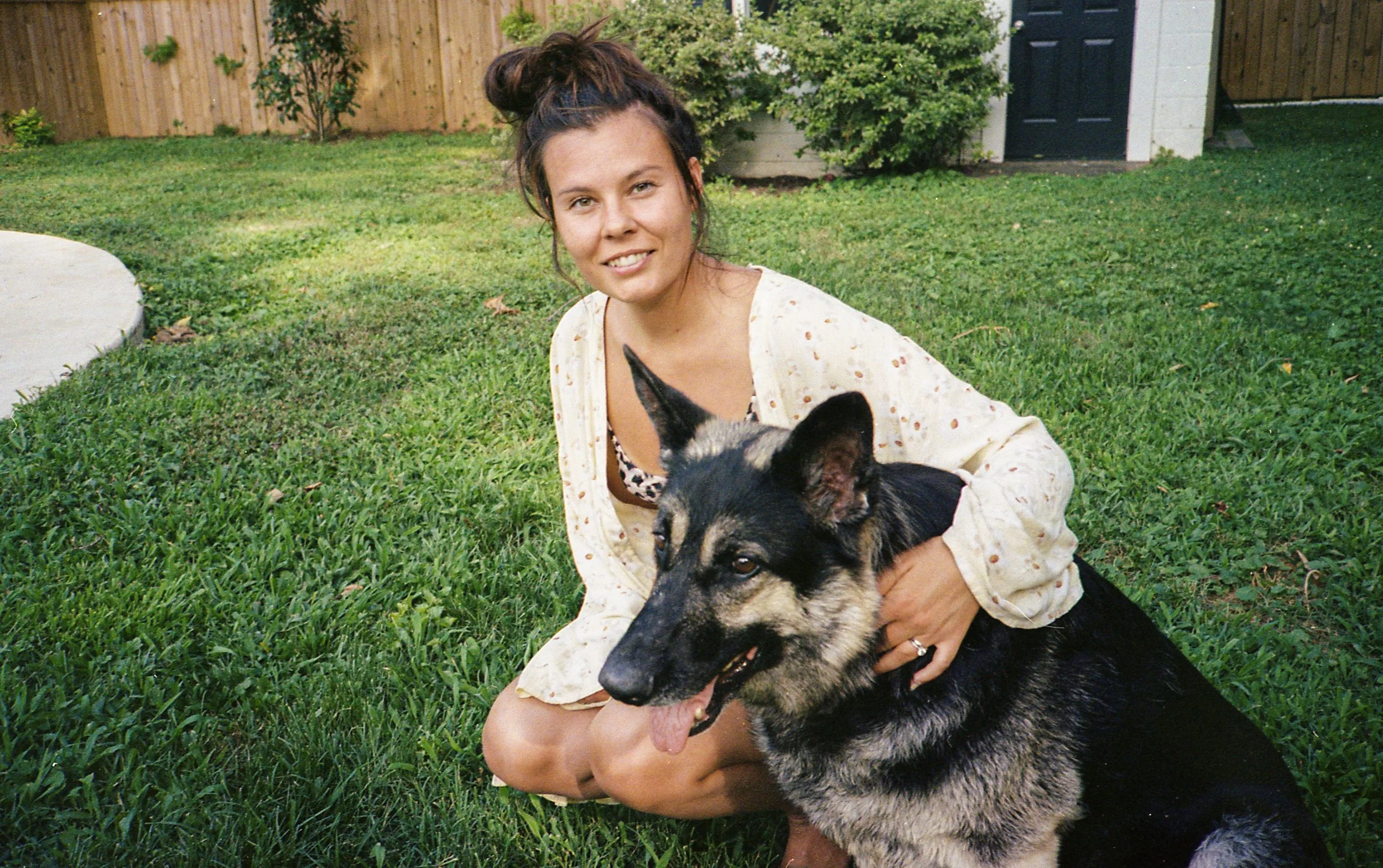 Una mujer con cabello recogido en un moño, vestida con una blusa beige y sentado en un césped, sosteniendo a un perro pastor alemán en un entorno de jardín con cercas y árboles.