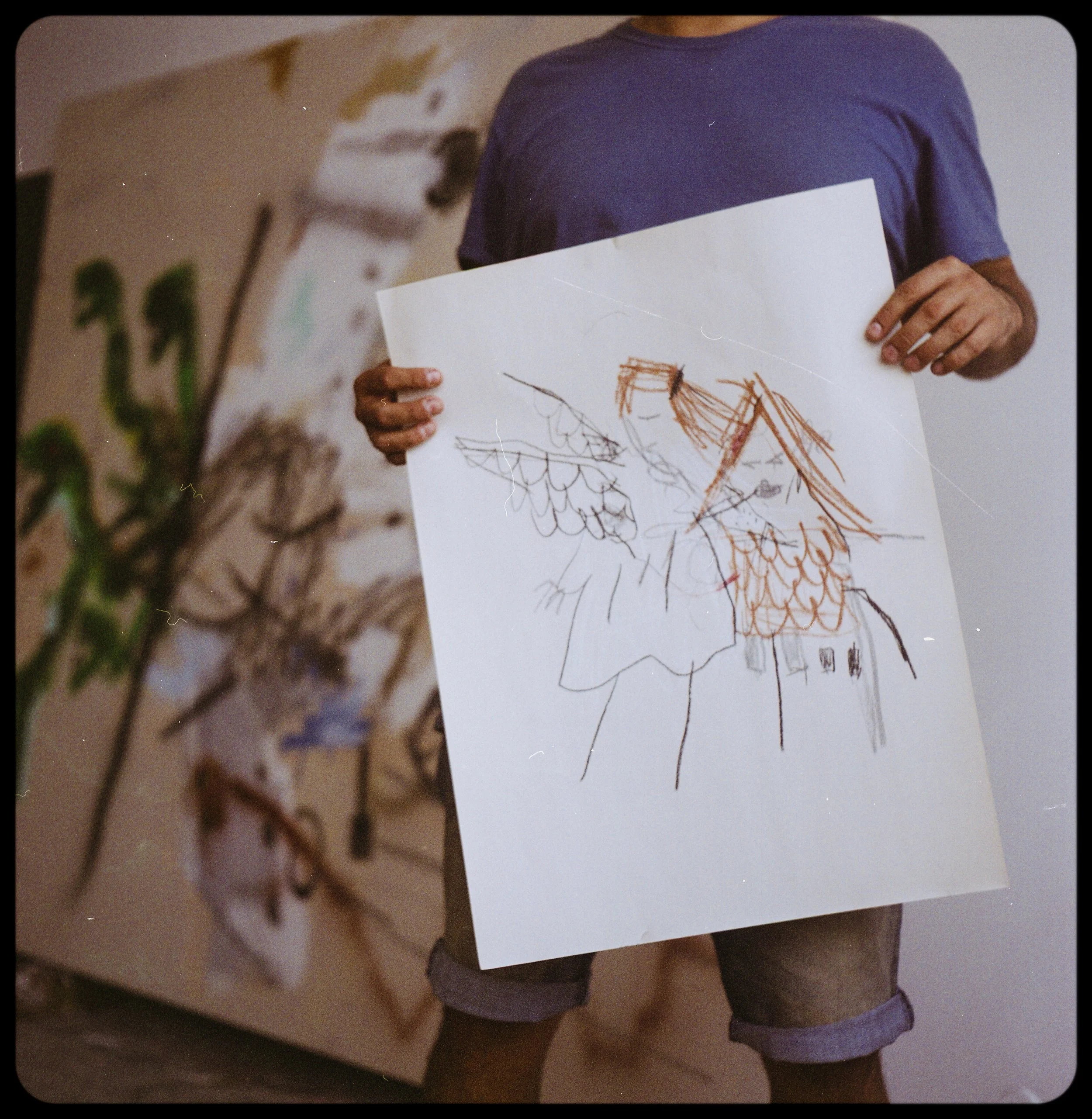 Persona sosteniendo un dibujo infantil de un ángel con alas y cabello largo, hecho con lápices y colores.