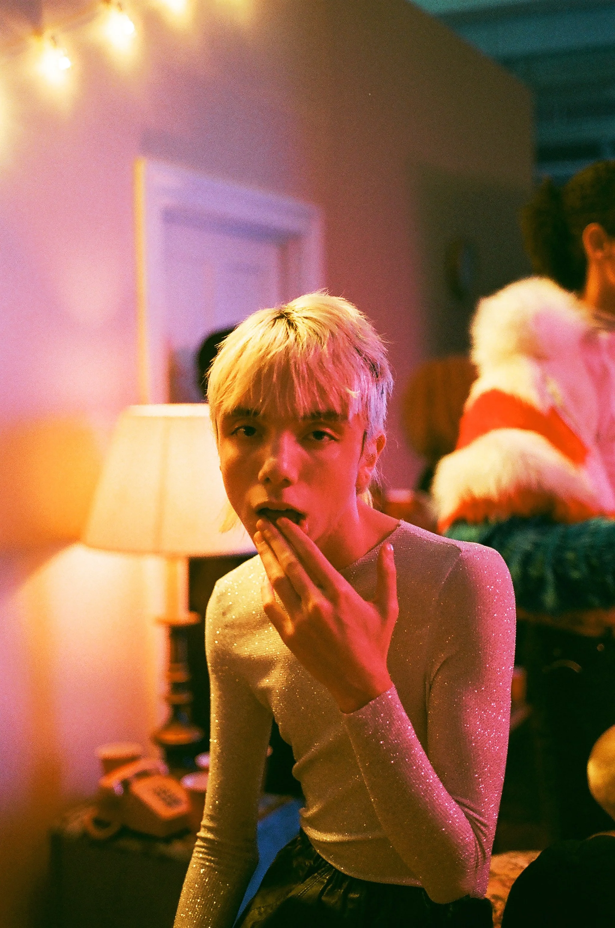 Joven con cabello rubio y camiseta brillante, en una fiesta con luces cálidas, con una mano en la boca, en un ambiente decorado con luces y gente al fondo.