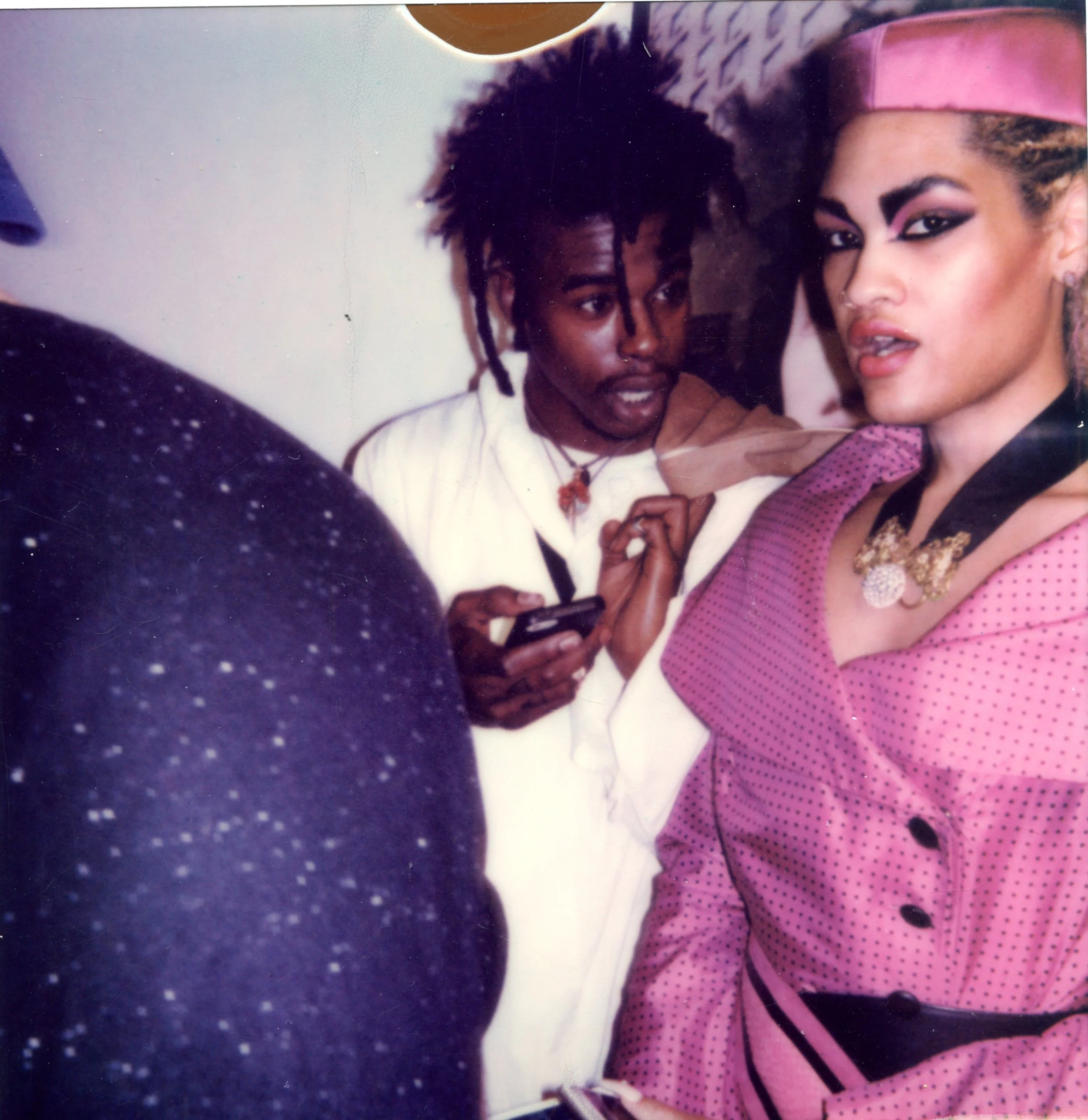 Una mujer con ropa rosa y maquillaje llamativo observa hacia la cámara mientras un hombre con peinados rasta revisa su teléfono móvil, en un entorno interior con fondo difuso.