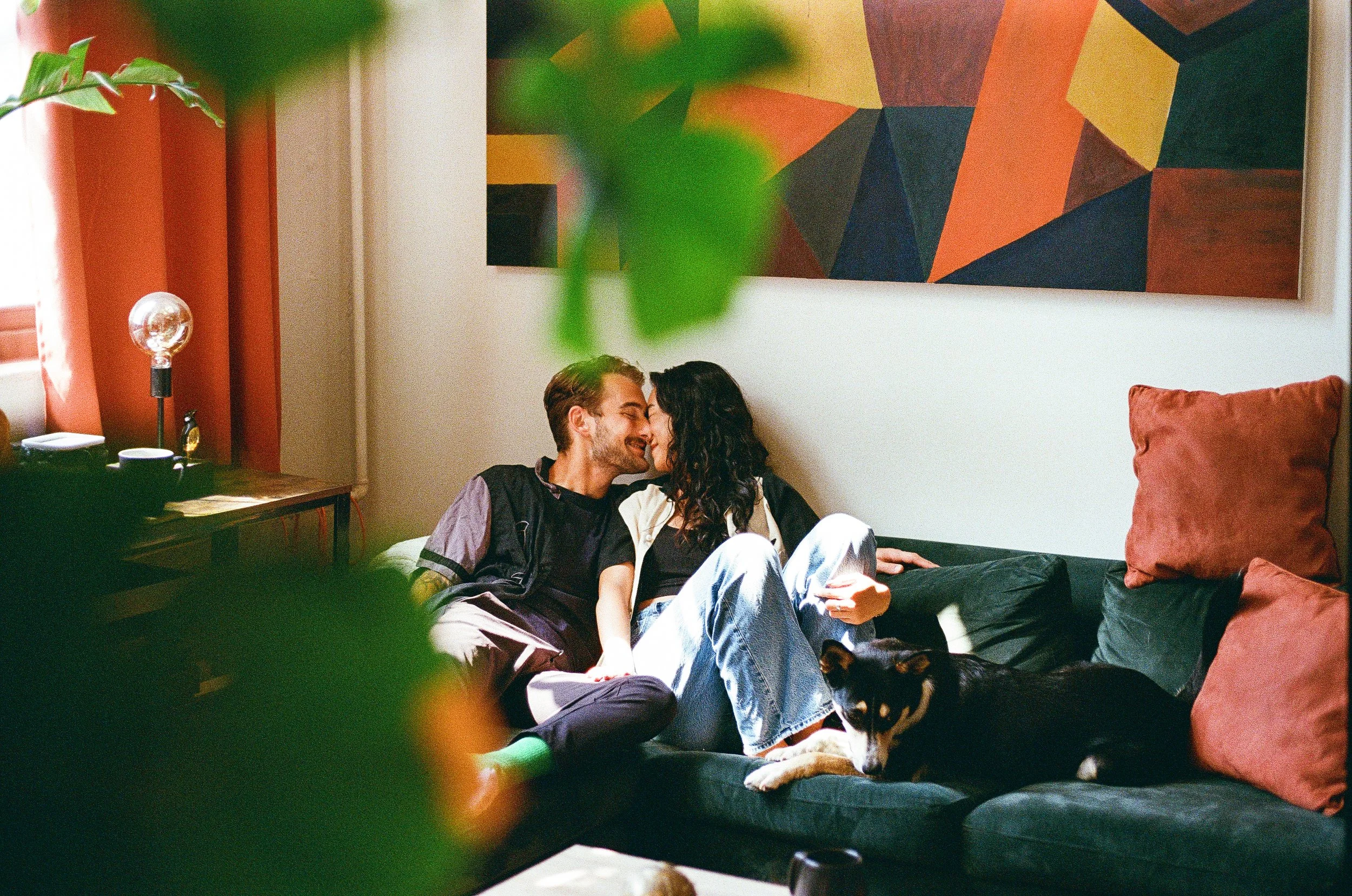 Una pareja riendo y abrazándose en un sillón, con un perro acostado en el mismo sillón, en una sala decorada con cuadros abstractos y cojines, con luz natural entrando por la ventana.