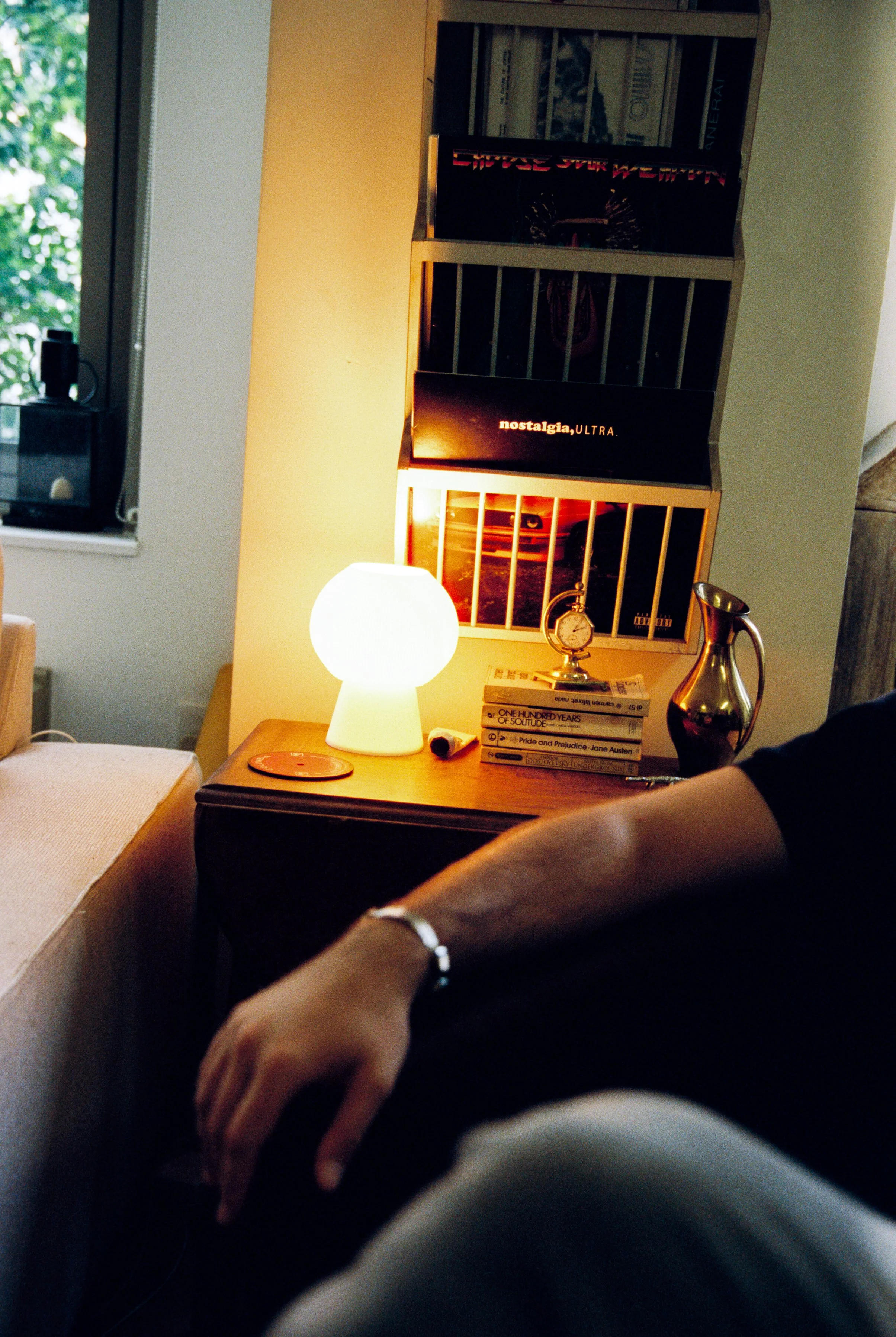 Una mesa junto a una pared con una lámpara de luz blanca, varios libros apilados, un reloj, un jarrón y un teléfono. En la pared hay varias portadas de vinilos y un libro titulado 'nostalgia, ULTRA'.