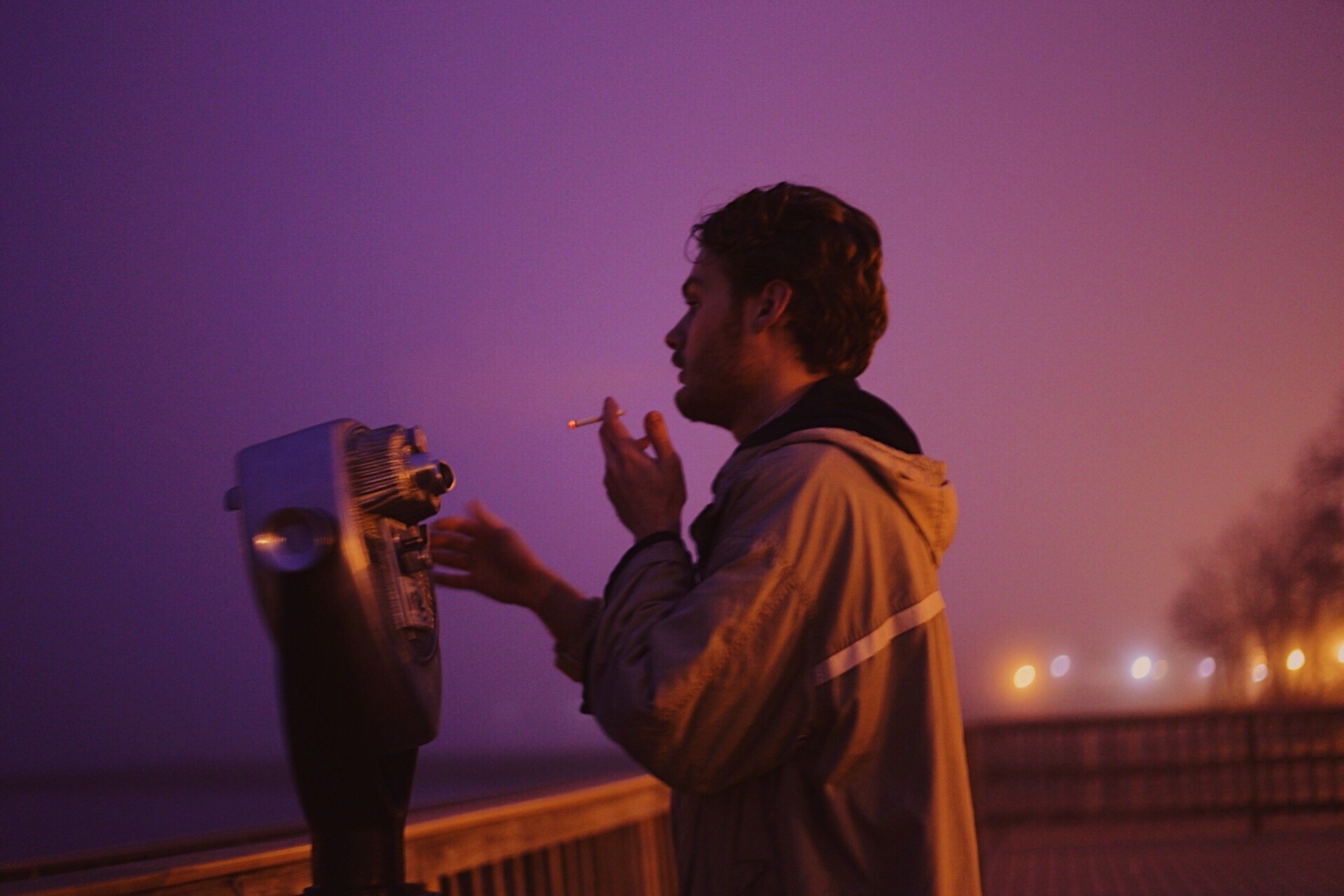 Joven hombre con chaqueta en la playa en la noche, usando un binocular y fumando con fondo de cielo morado y farolas iluminando
