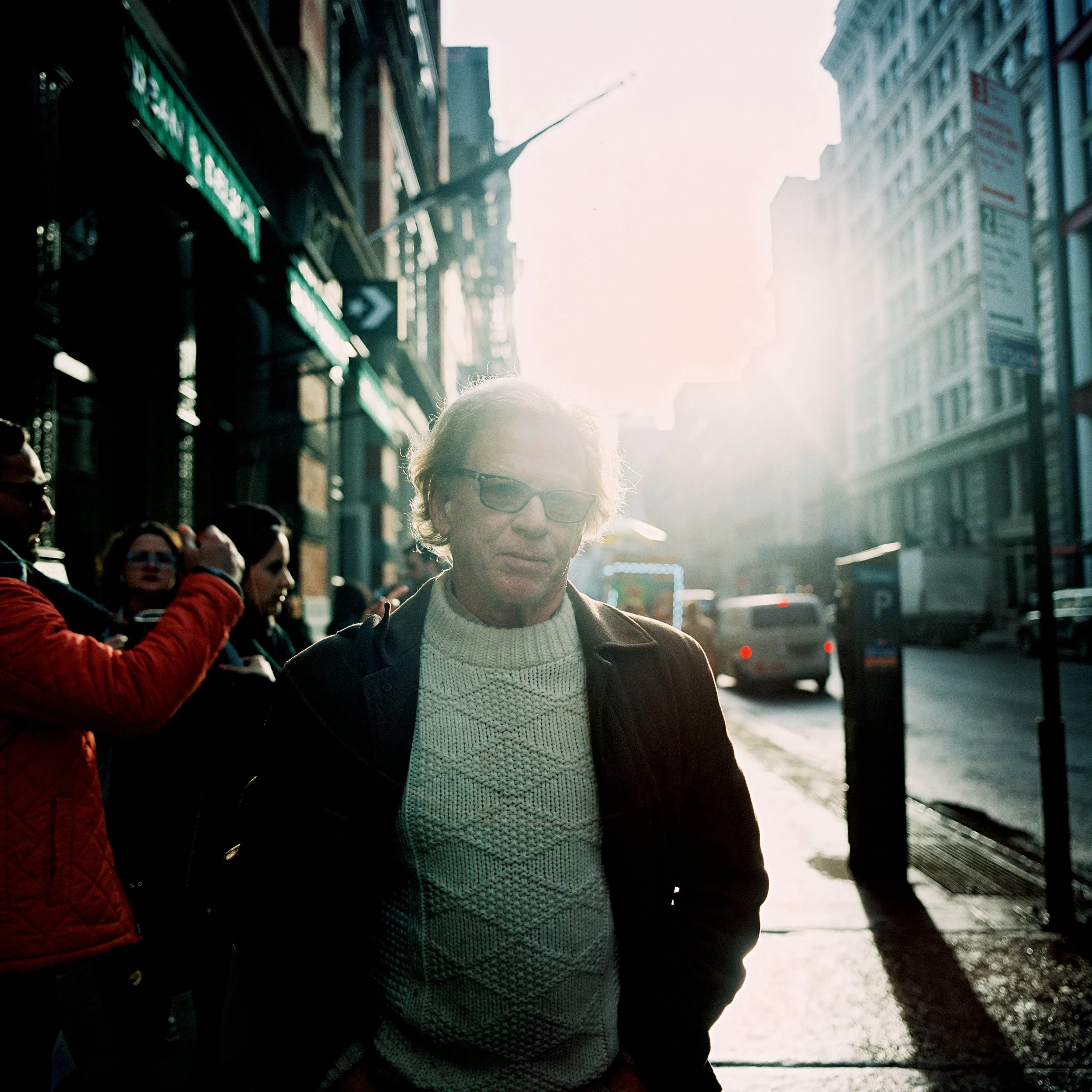 Hombre con gafas y suéter beige en la calle con tráfico y edificios altos, luz solar intensa de fondo