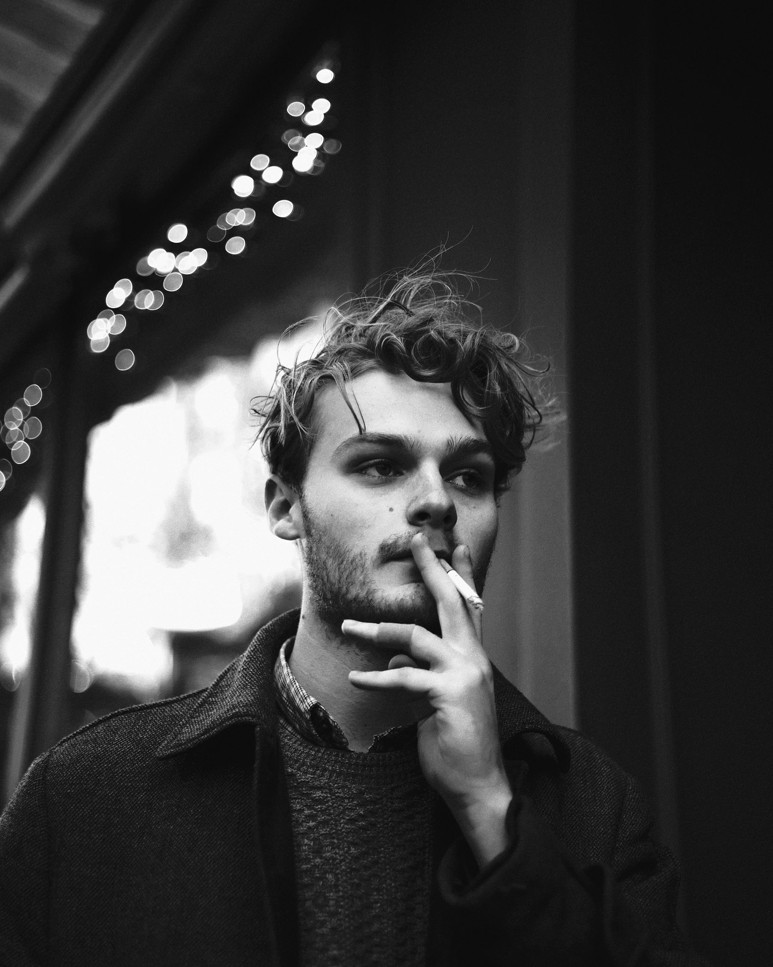 Joven sosteniendo cigarrillo en mano, viendo pensativo, en un entorno con decoraciones de luces en el fondo, en blanco y negro.