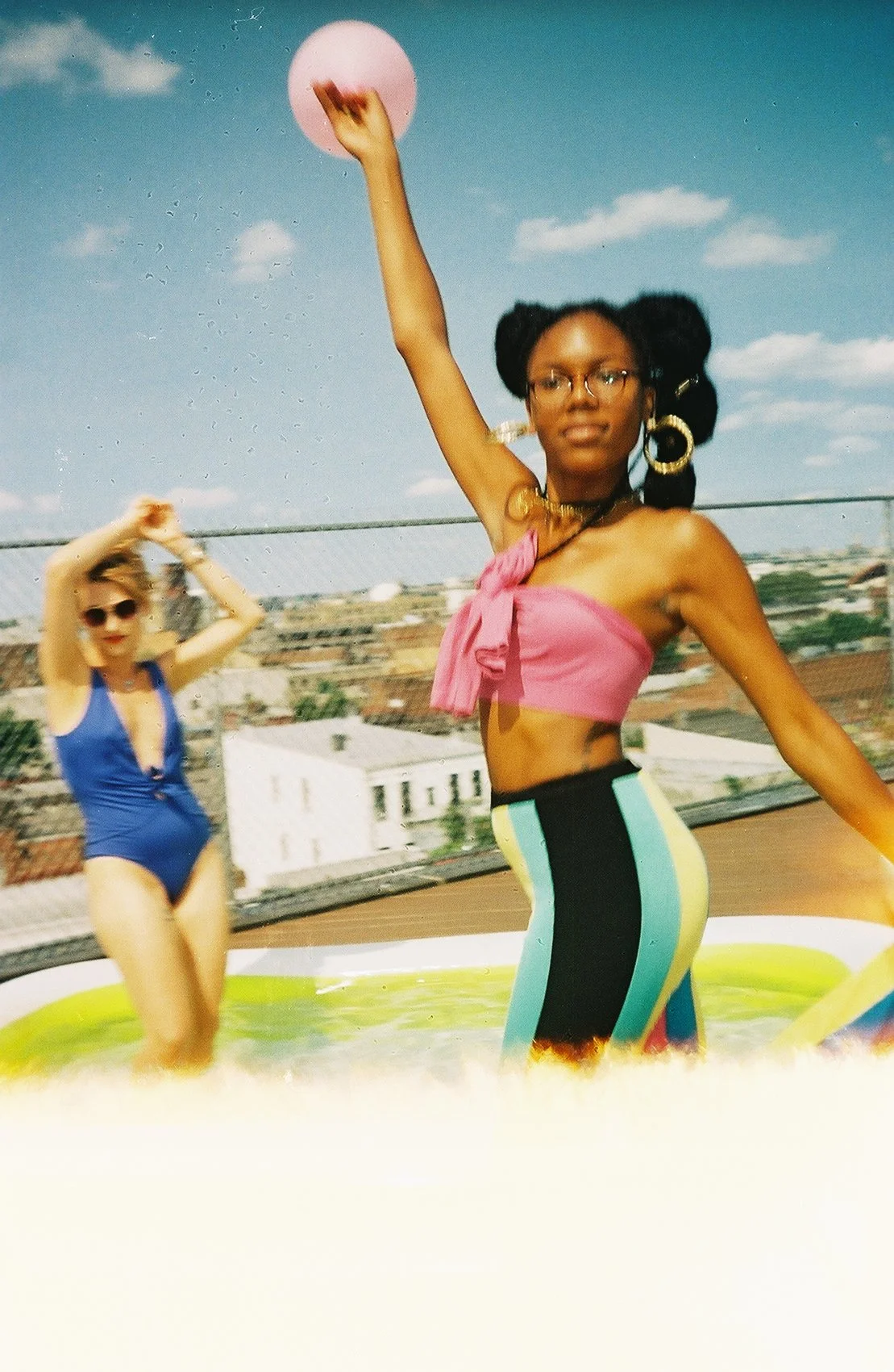 Dos mujeres en una piscina en la azotea, una con un vestido azul y la otra con un top rosa y pantalones color block, levantando los brazos bajo un cielo azul con algunas nubes.