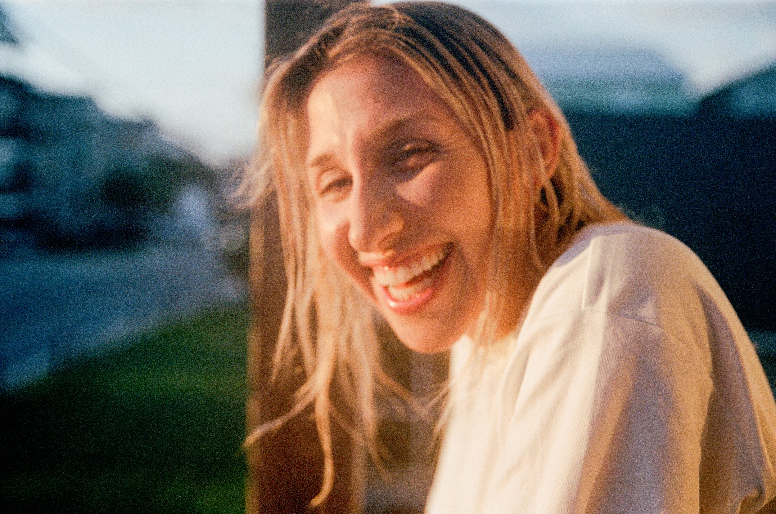 Joven mujer sonriendo al sol, con cabello rubio y rizado, vestida con camiseta blanca, en un entorno exterior durante el atardecer.